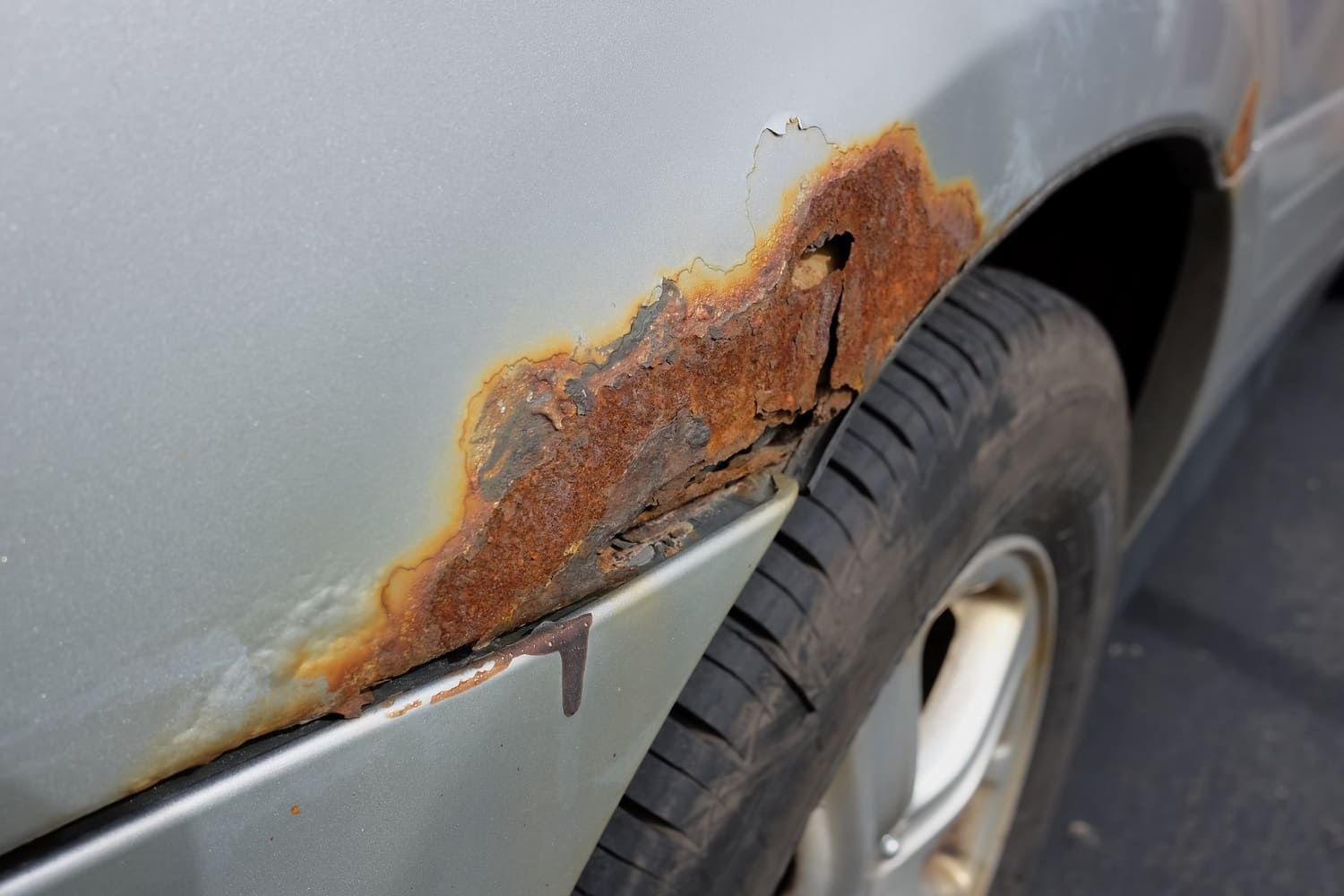 Close-up of a rusty and corroded area on a silver car's body near the wheel well.