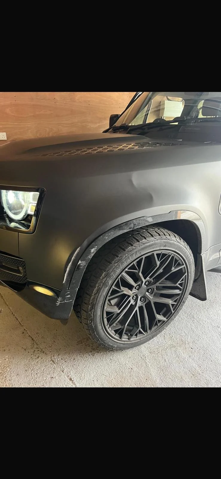 Close-up of a black luxury SUV with a damaged front fender and scratched wheel, parked indoors next to a wooden wall.