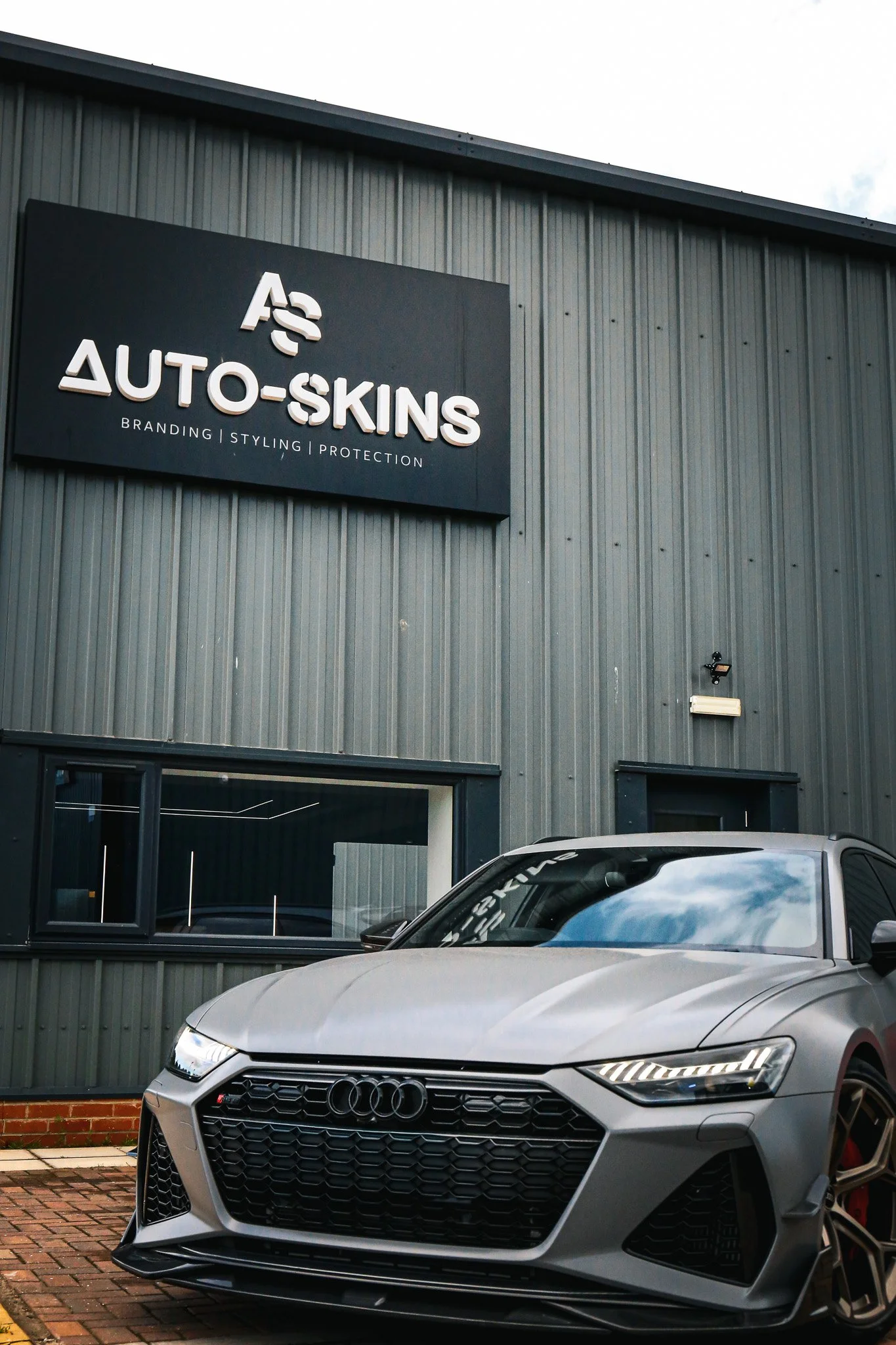 A silver Audi RS vehicle parked in front of a gray building with a sign that reads 'Auto-Skins' and offers branding, styling, and protection services.