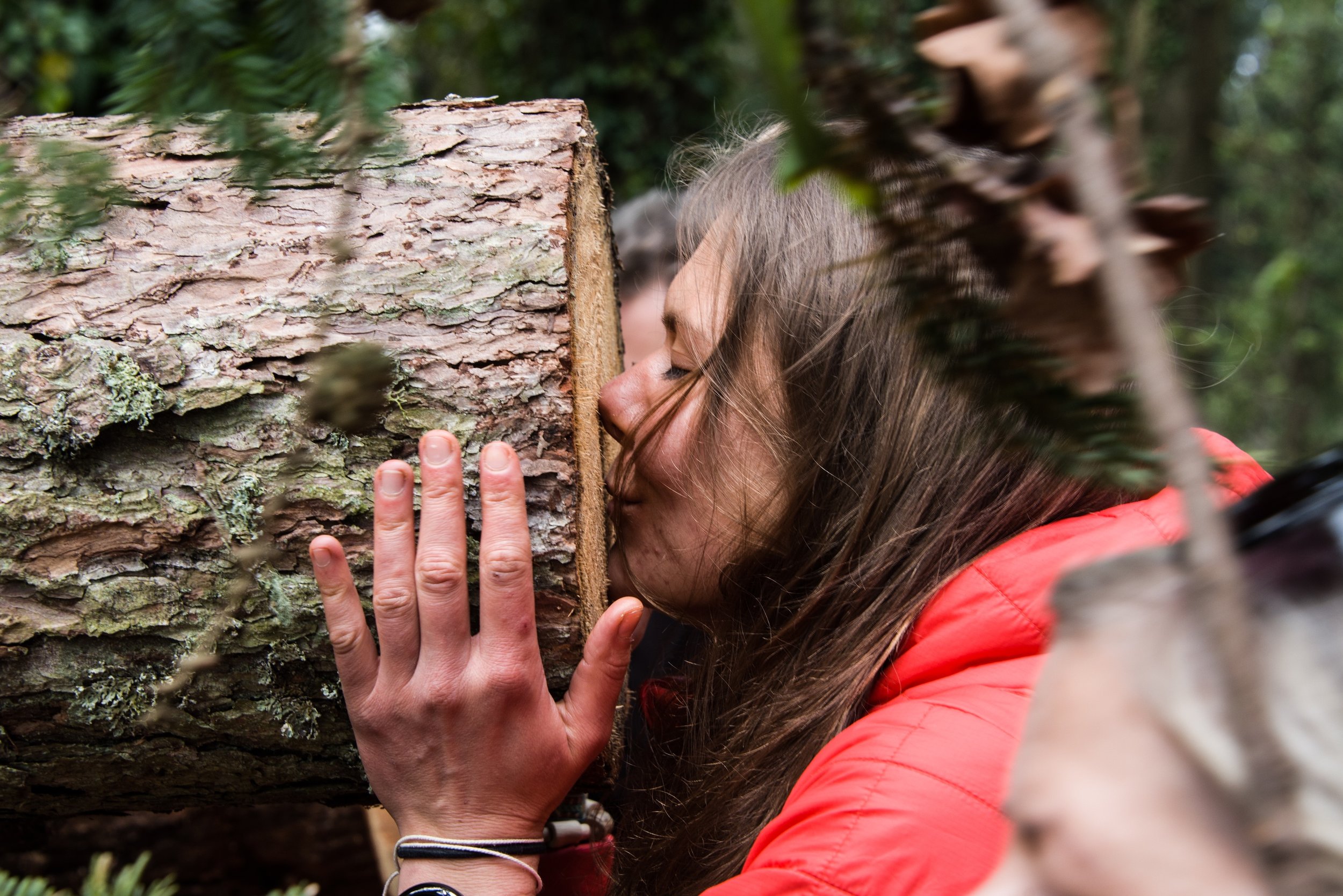 Lady at Go Wild Education Forest School training, with long brown hair has her face against the cut end of a larch log. She has her hands around the log and her eyes are closed. She is enjoying the scent of the wood.