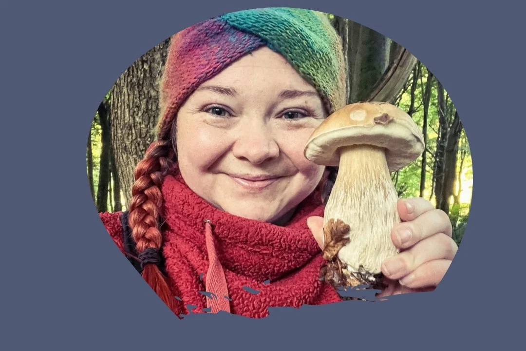 A woman smiling while holding a large mushroom outdoors in a wooded area, wearing a colorful knit hat and red scarf.