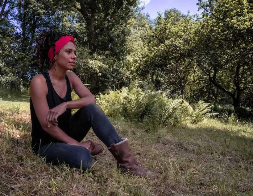 A woman with dark curly hair wearing a red headband and black sleeveless top sitting outdoors on a grassy area with trees in the background.