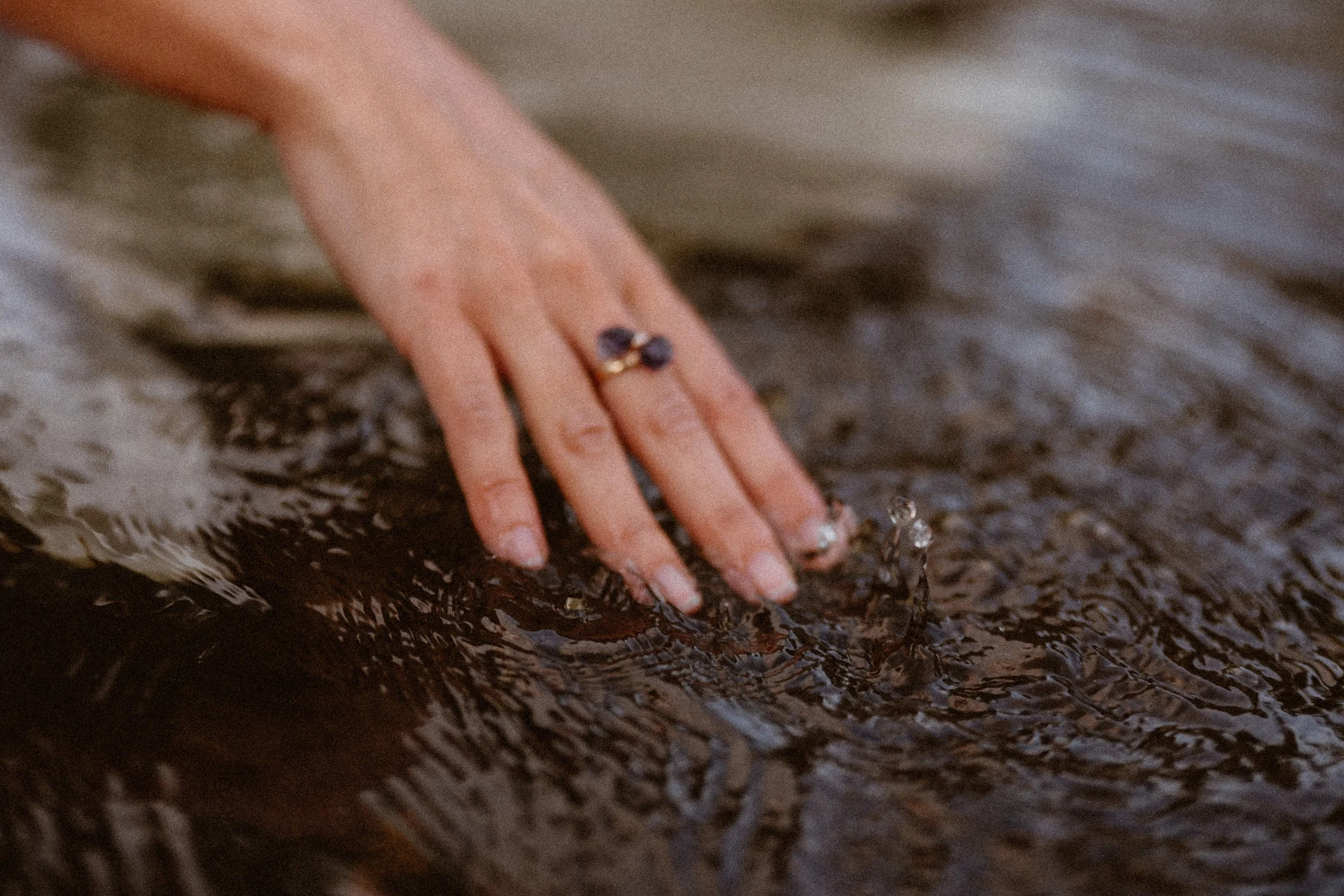 A hand wearing a purple stone ring with fingertips submerged in moving water.