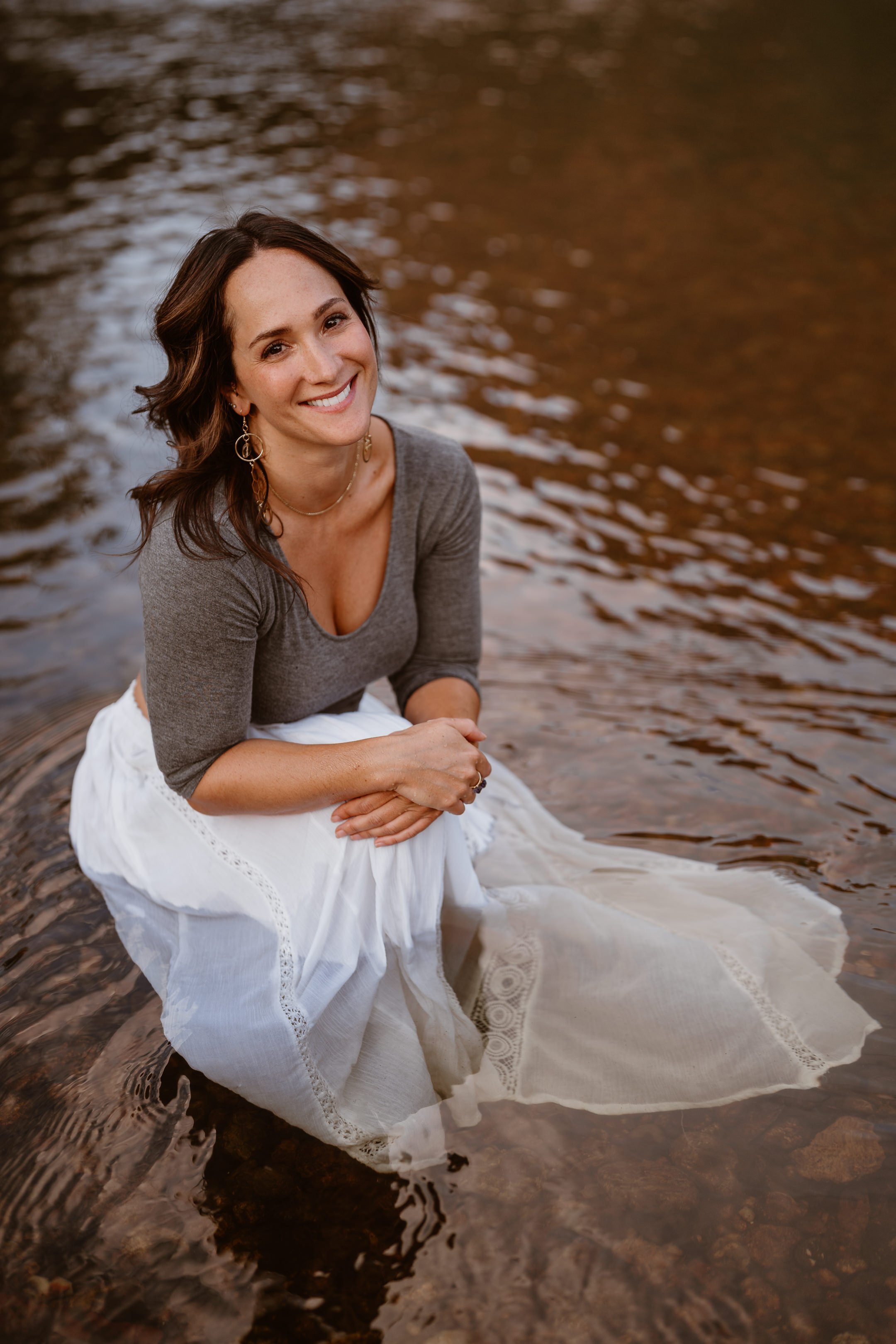Dori Lewis crouches smiling, her feet and white skirt in the river.