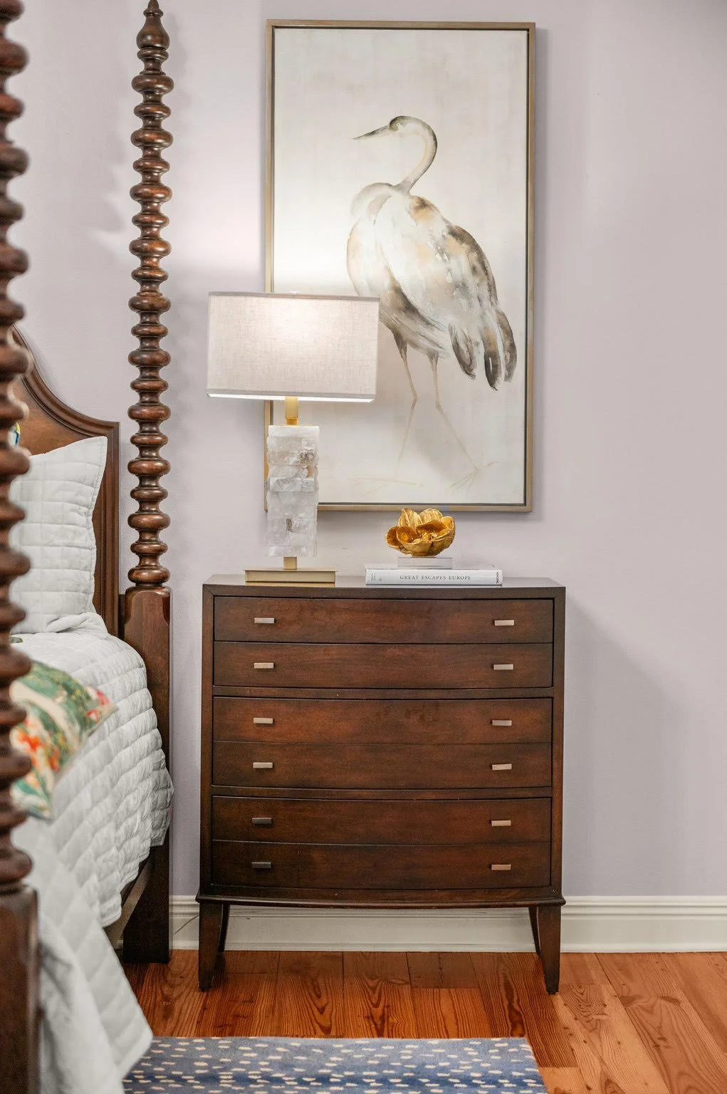 Ornate wooden bedframe and nightstand with marble based lamp and painting of an egret