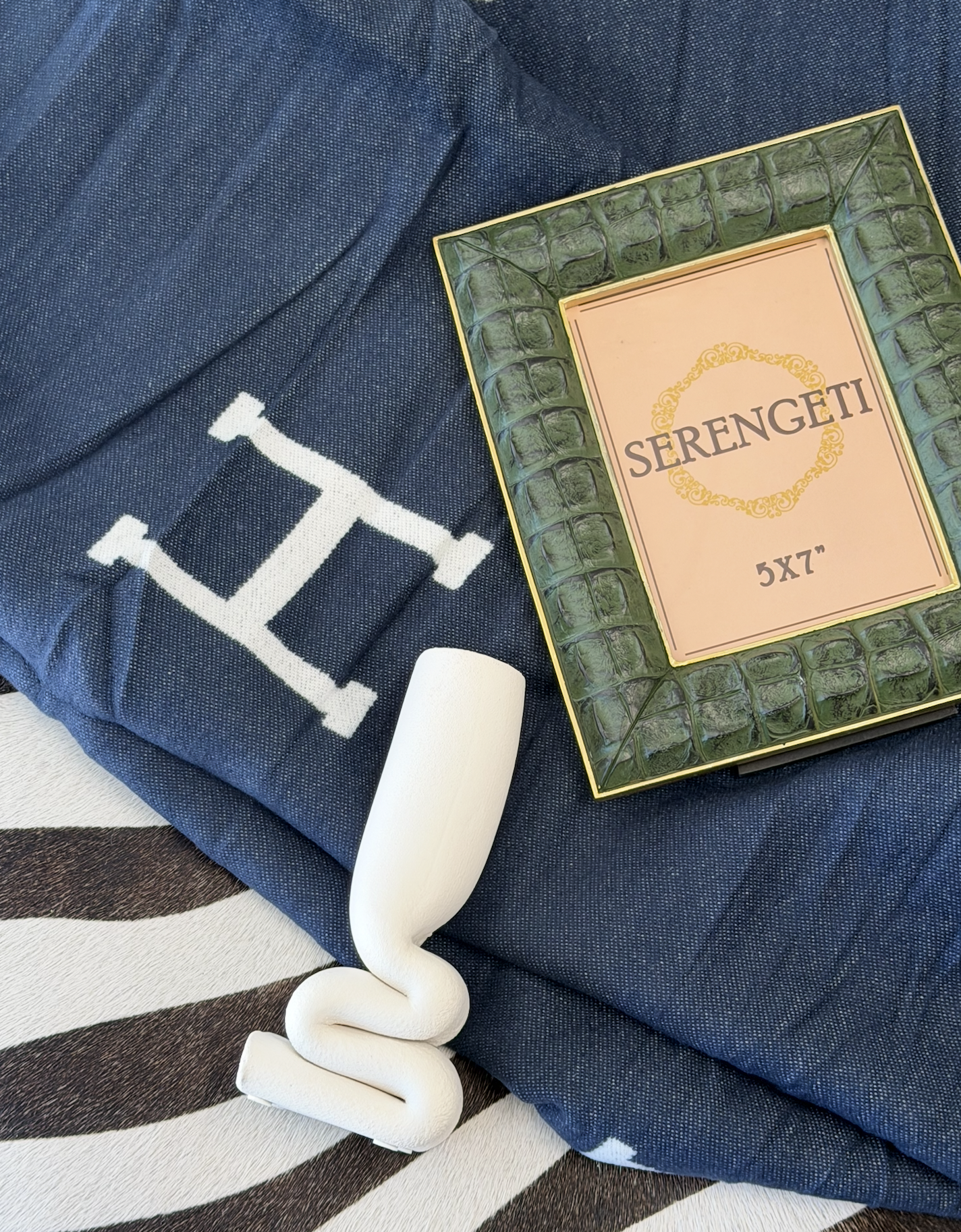 Crocodile green picture frame, squished white bud vase, H logo blanket on top of a zebra ottoman