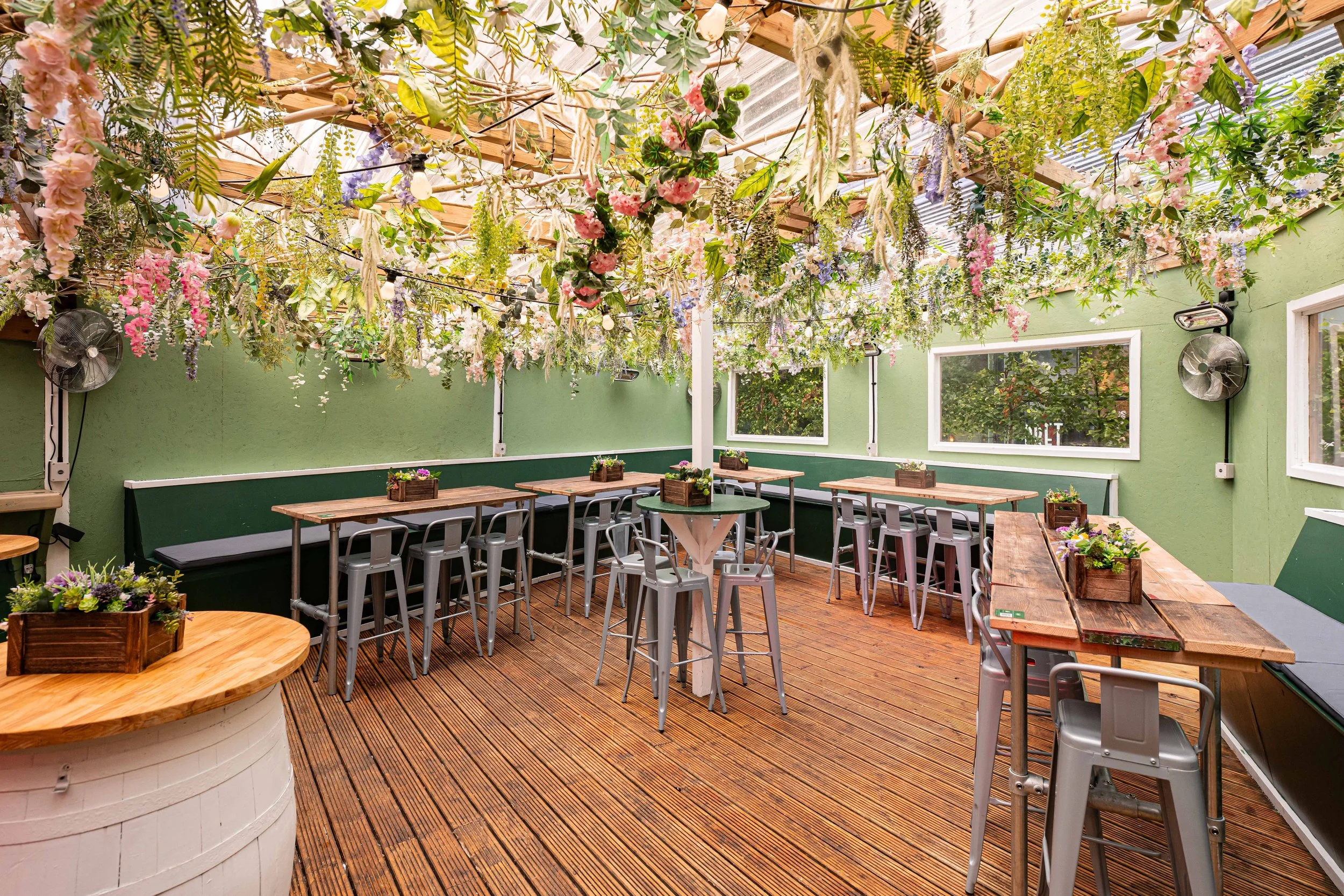 Indoor patio dining area with wood flooring, green walls, and large windows. Hanging floral decorations above fixtures and several tables with chairs and flower boxes.
