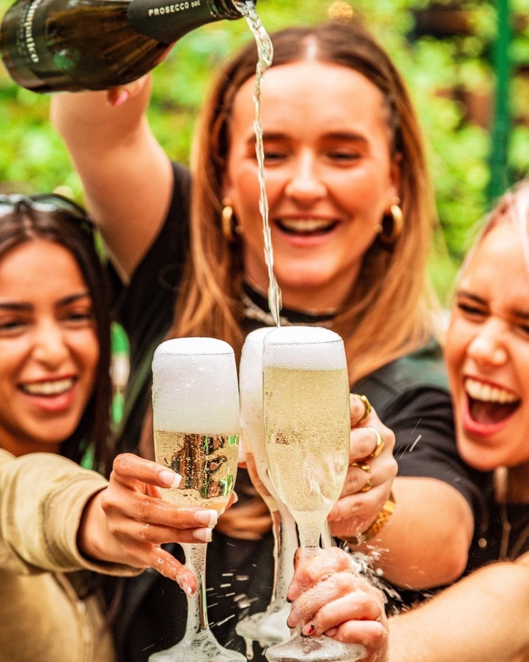 Three women celebrating with champagne outdoors, smiling, as one pours Prosecco into glasses.