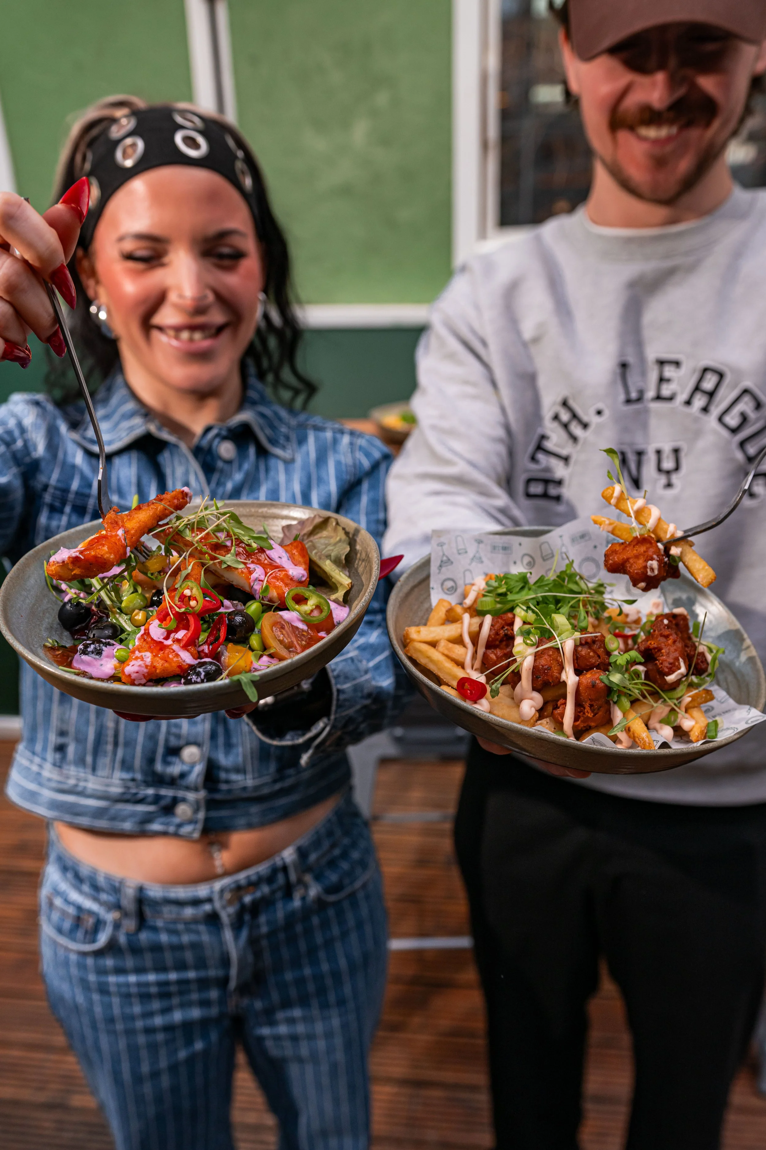 Two people holding plates of colorful salads and fries, smiling indoors.
