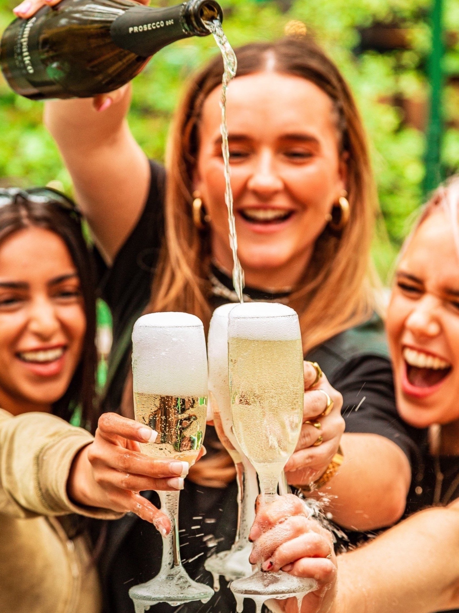 A group of friends celebrating and toasting with glasses of champagne, with one woman pouring champagne from a bottle labeled 'Prosecco DOC'.