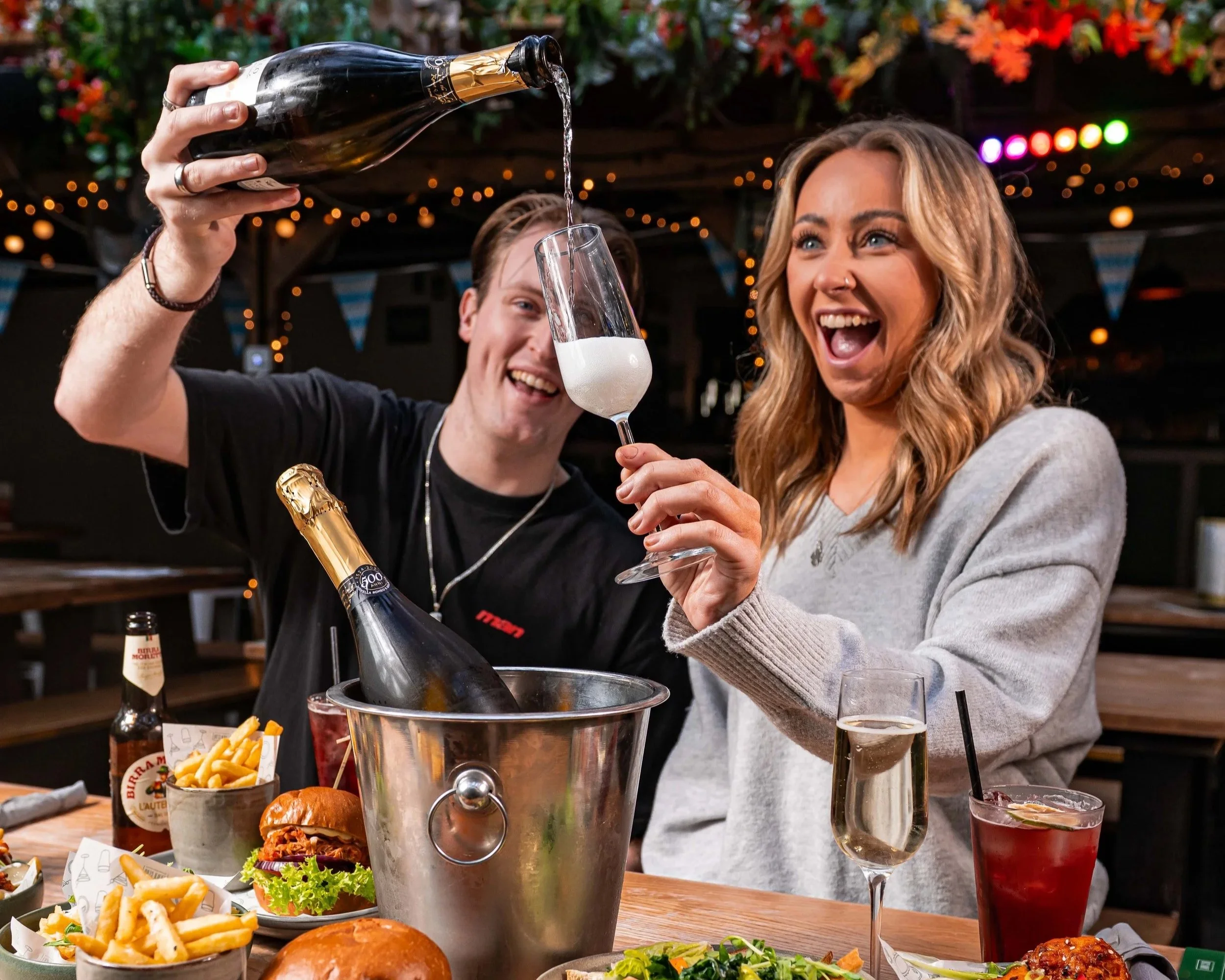A man and woman celebrating at a restaurant, pouring champagne into a glass, surrounded by food and drinks, with decorative string lights in the background.
