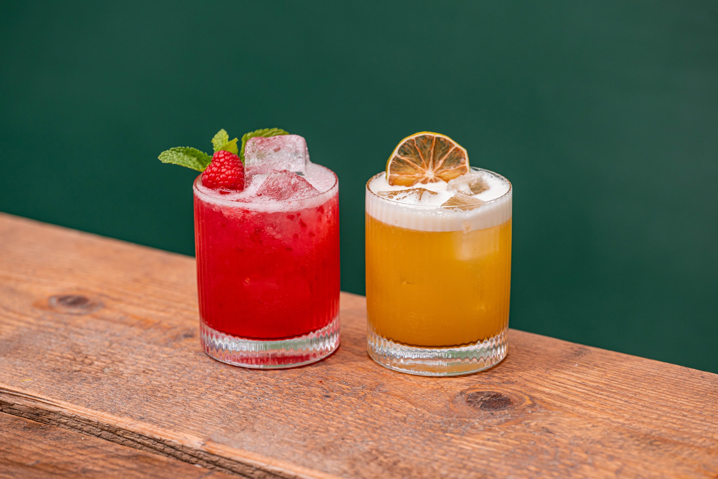 Two colorful cocktails, one red garnished with a raspberry and mint, the other yellow garnished with a dried lemon slice, on a wooden surface against a green background.