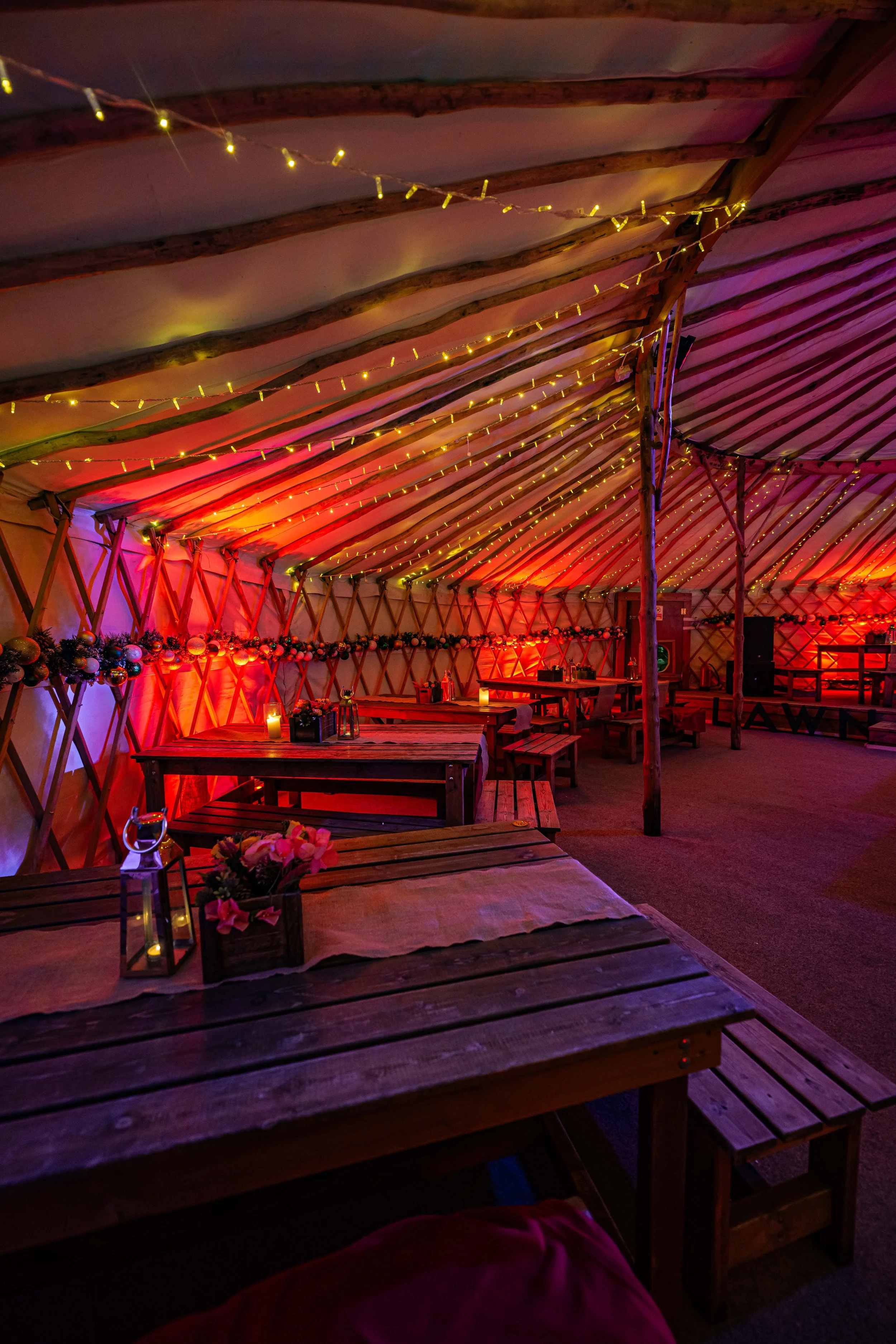 Inside a decorated event yurt tent with wooden tables adorned with flowers, lanterns, and candles. The ceiling features string lights and wooden beams, with warm coloring and festive decorations.