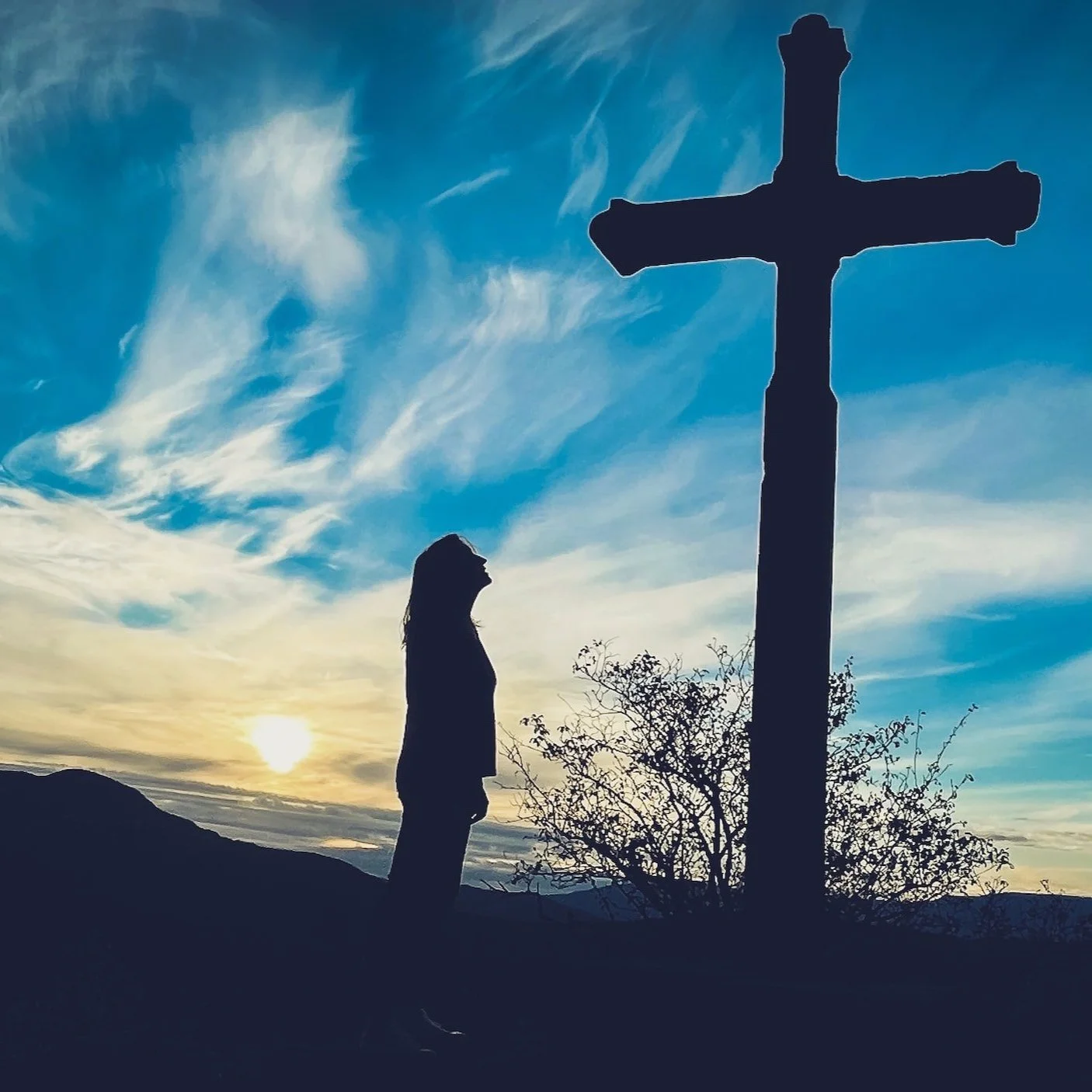 Silhouette of a person standing beside a large cross on a hill with a dramatic sky at sunset.