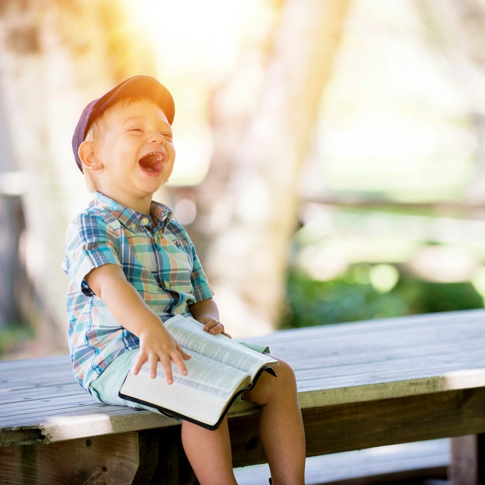Young boy laughing, sitting on a bench outdoors, holding a bible.