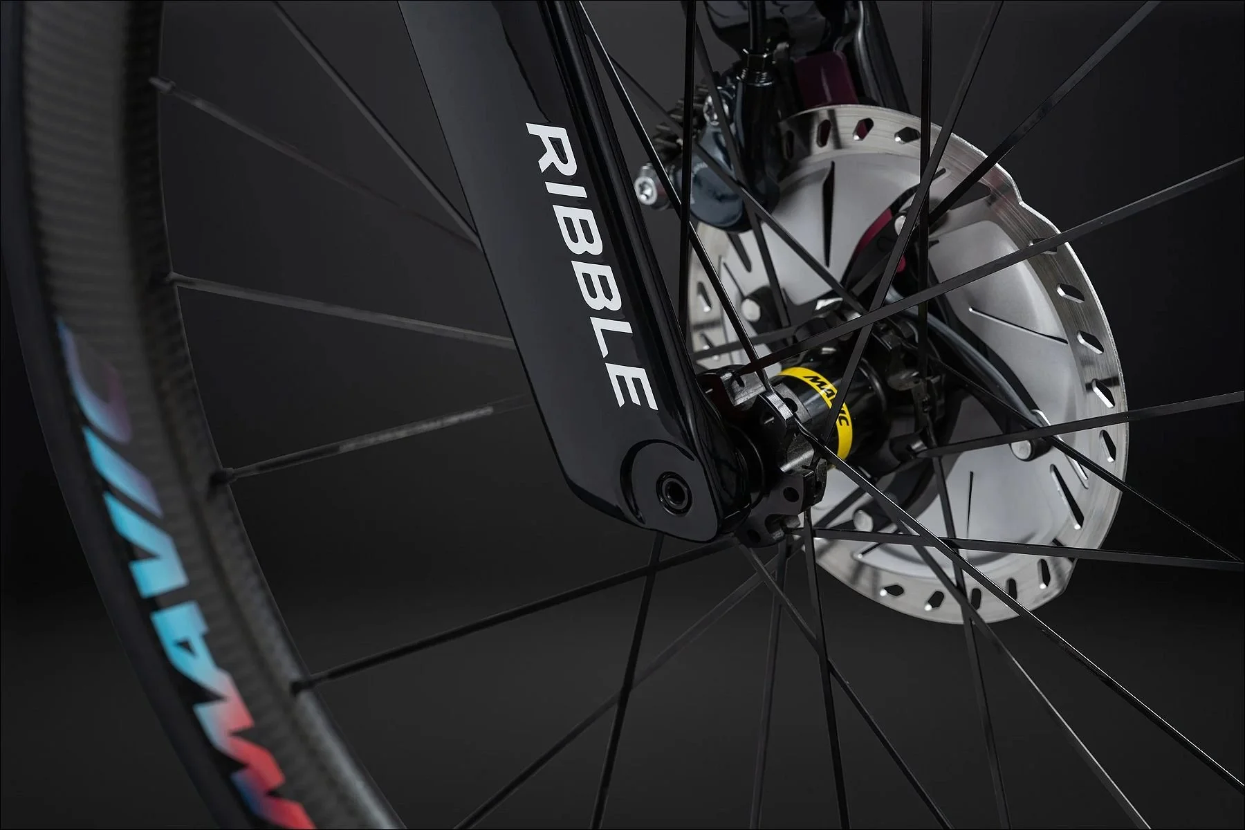 Close-up of a bicycle disc brake and wheel with Ribble branding on the fork against a dark studio background.
