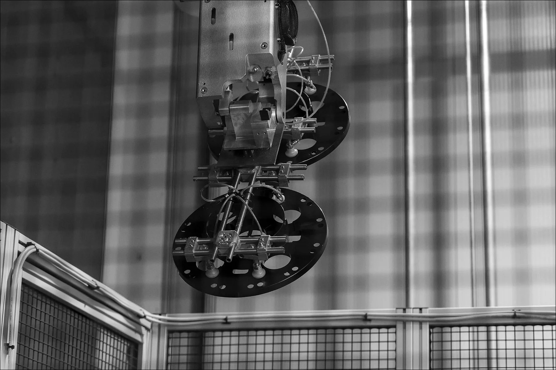 Black-and-white image of a suspended industrial robotic apparatus above a railing in a manufacturing facility.
