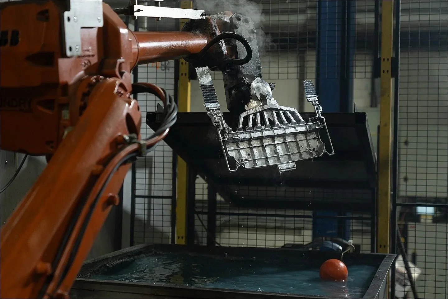 Industrial robotic arm lifting a metallic component inside a caged manufacturing cell.