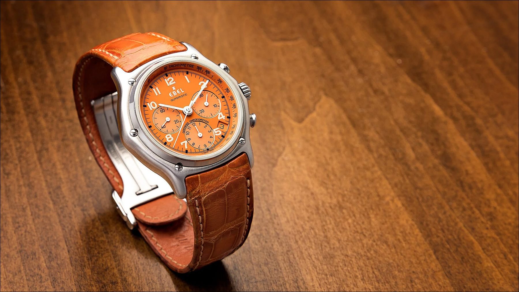 Orange dial wristwatch with a brown leather strap and polished steel case displayed on a wooden surface.
