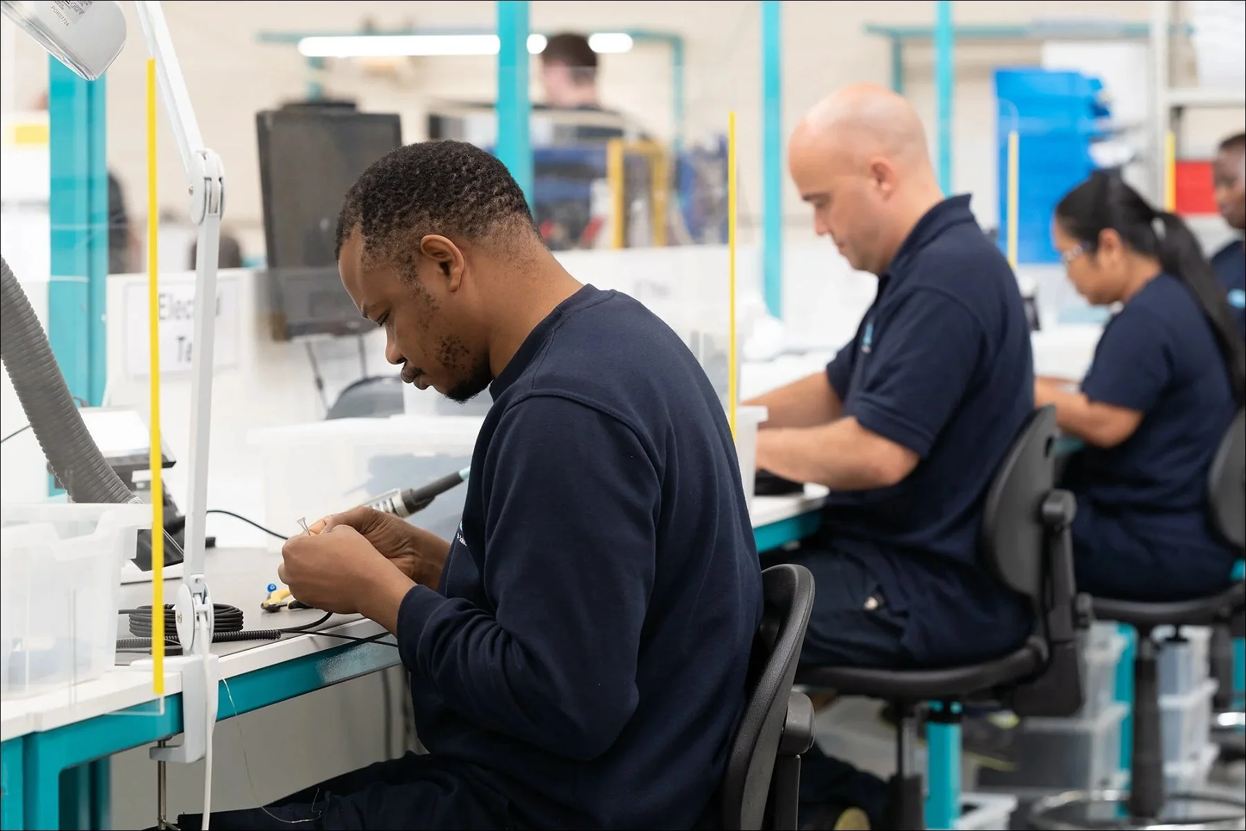 Two factory workers assembling components at an assembly line, sitting at workstations with tools.