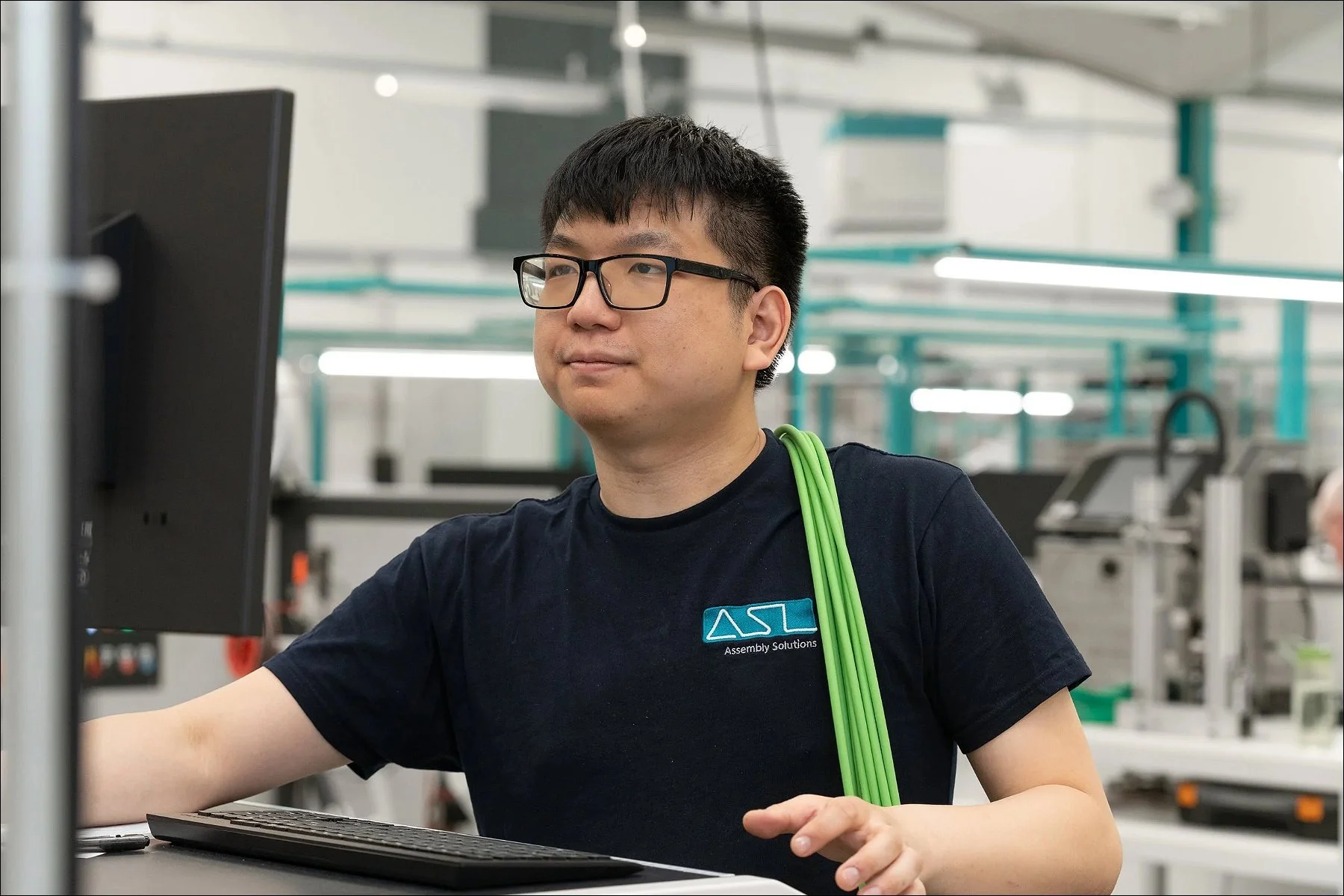 Technician working at a computer workstation in a manufacturing facility, wearing a dark uniform and glasses.