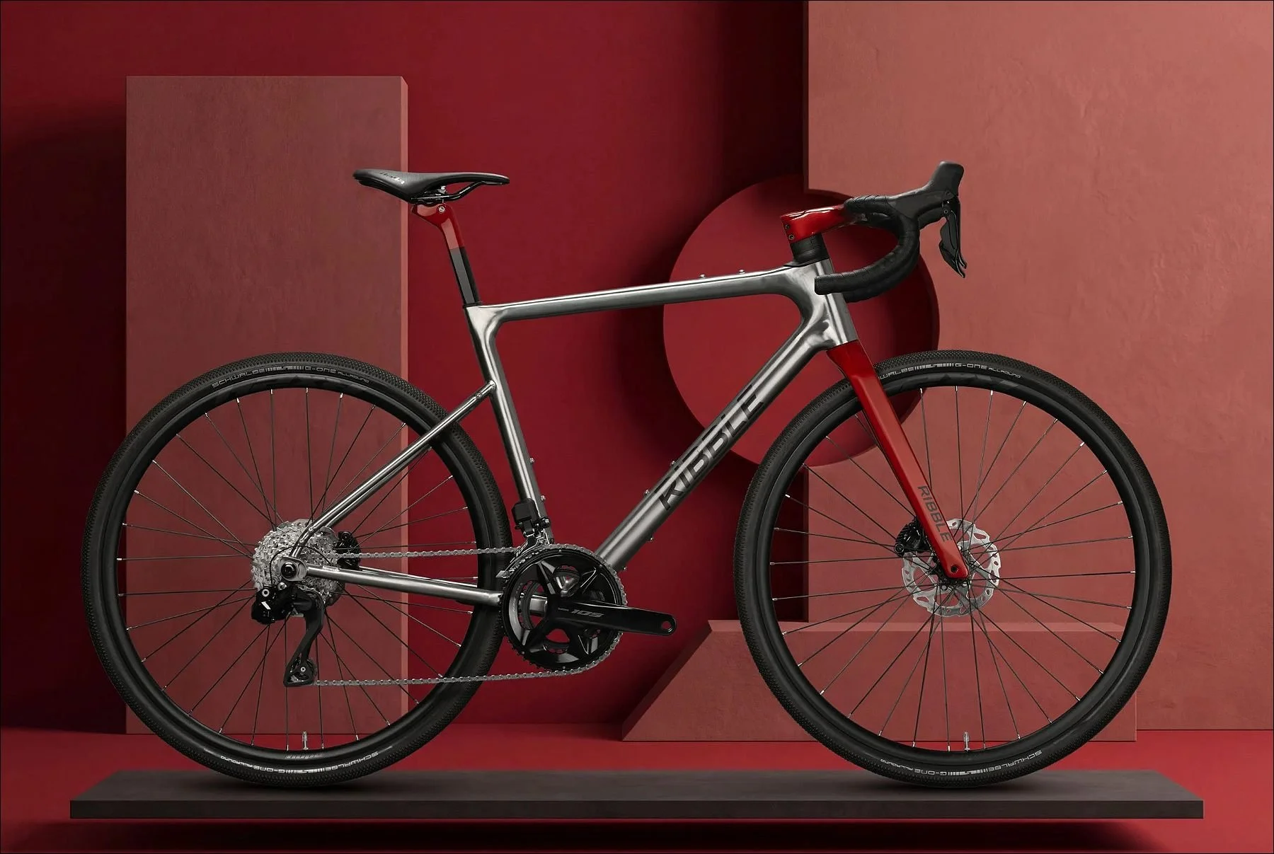 Silver road bicycle with red handlebar and wheels displayed on a two-level platform against a red and beige studio backdrop.