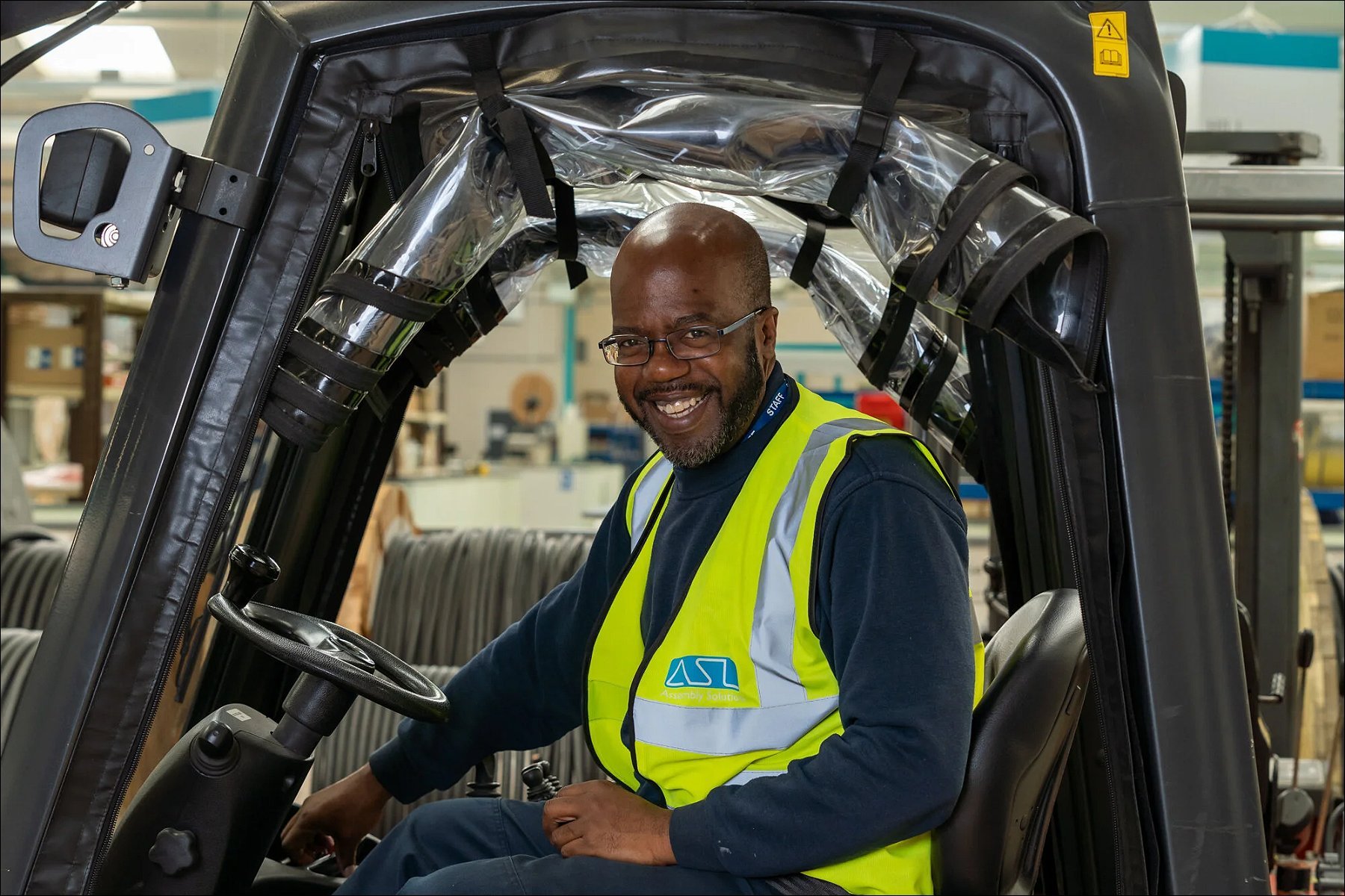 Factory worker wearing a high-visibility vest seated inside an industrial vehicle cabin and smiling at the camera.
