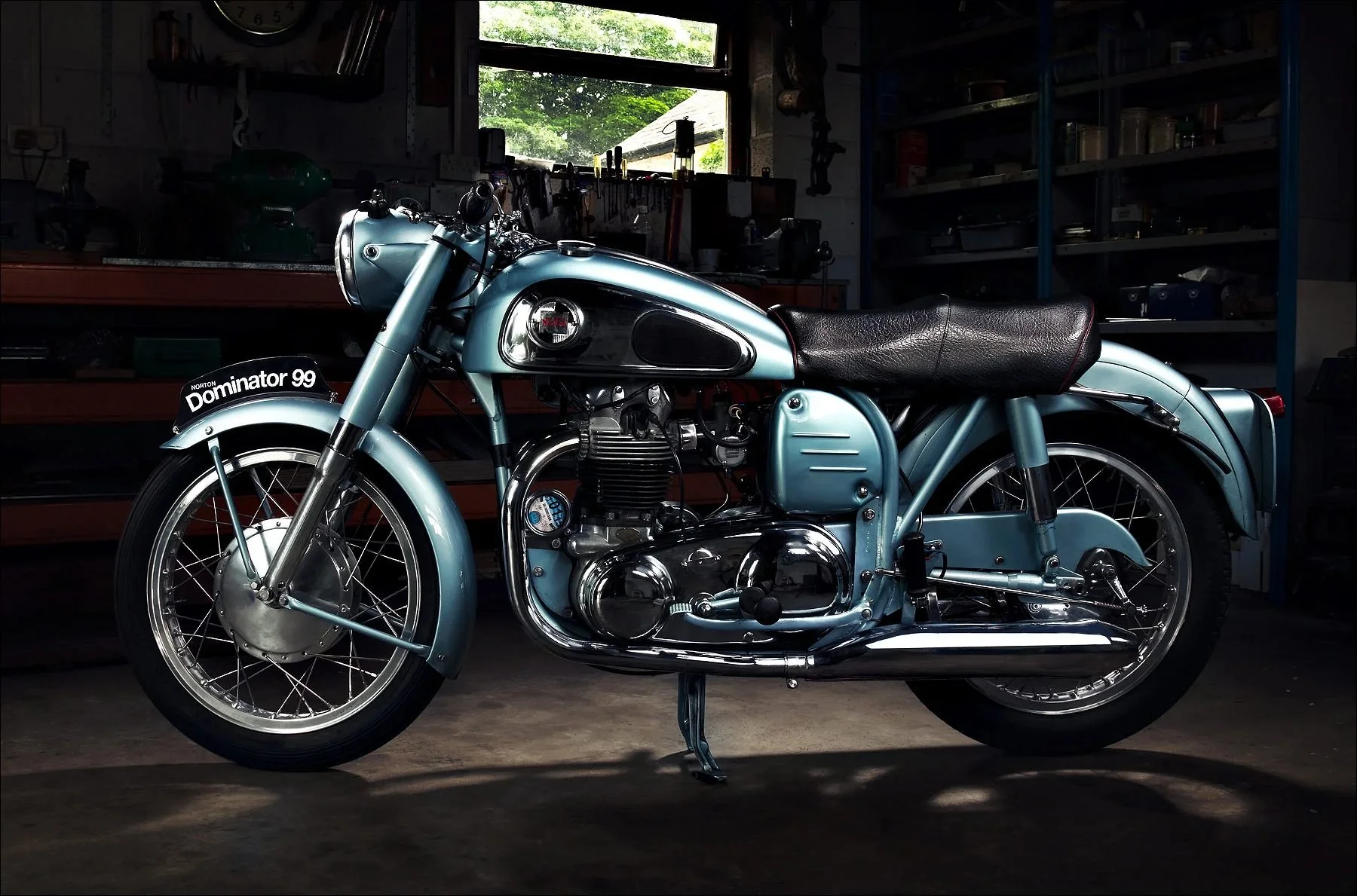Vintage blue motorcycle parked in a dimly lit workshop with various tools and equipment in the background