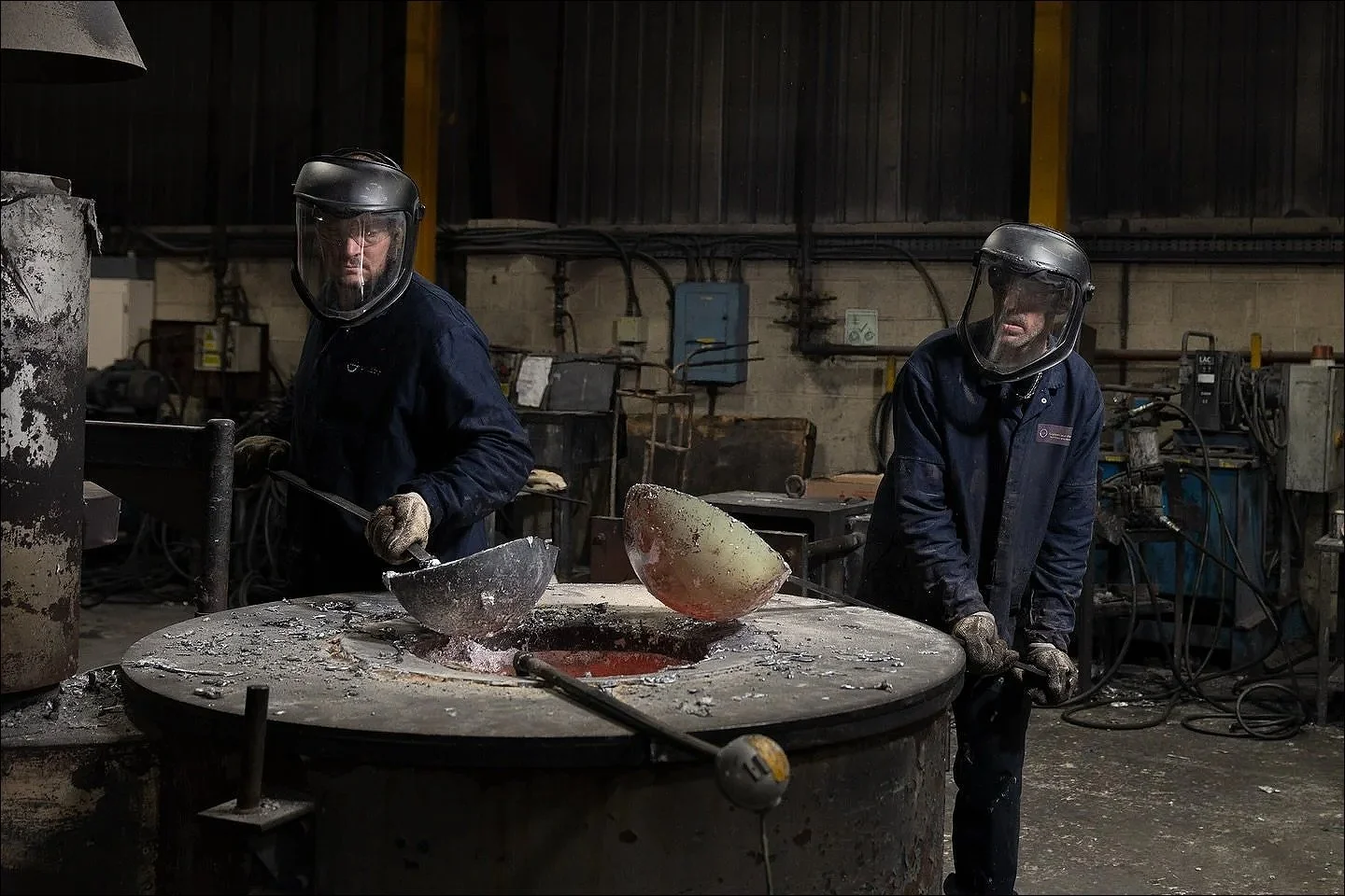 Two foundry workers wearing protective face shields standing next to a large casting table with molten metal.