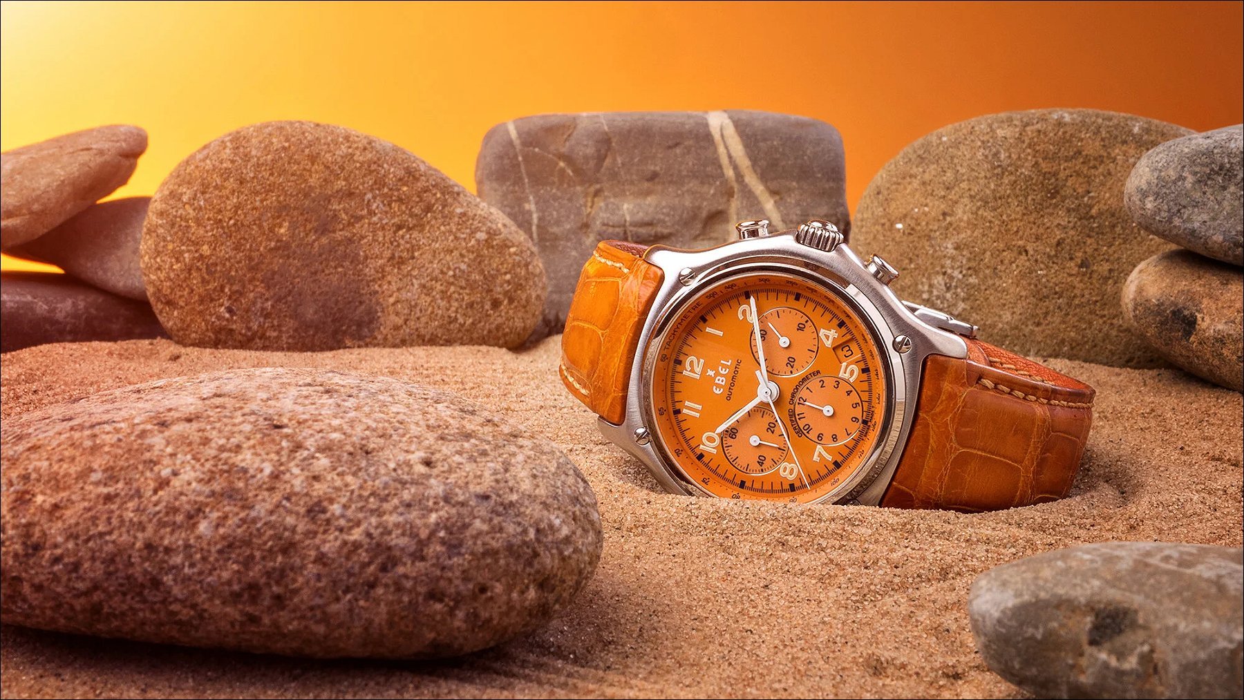 Orange watch with a brown leather strap displayed among smooth stones against a warm orange backdrop