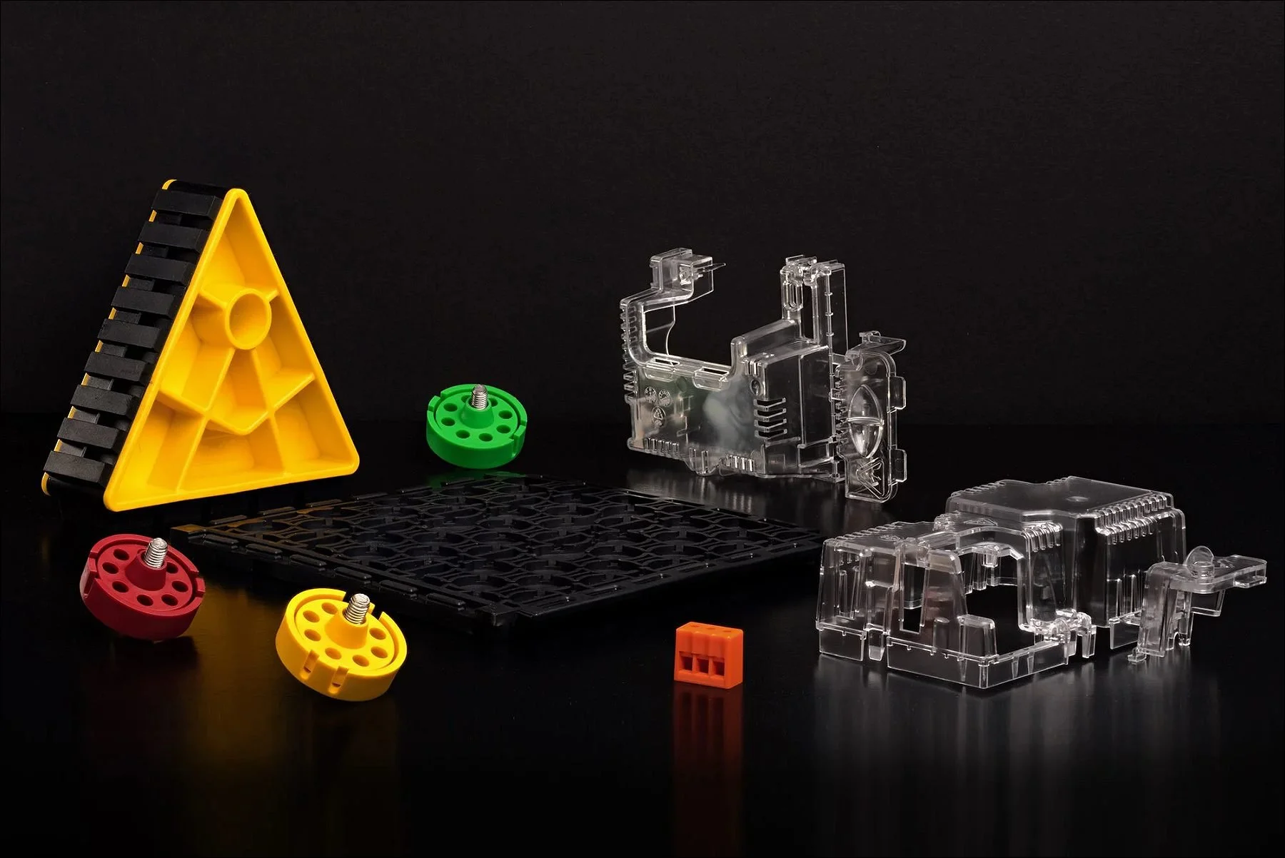 Assorted transparent mechanical components and a yellow triangular block with small colored cog pieces arranged on a black surface