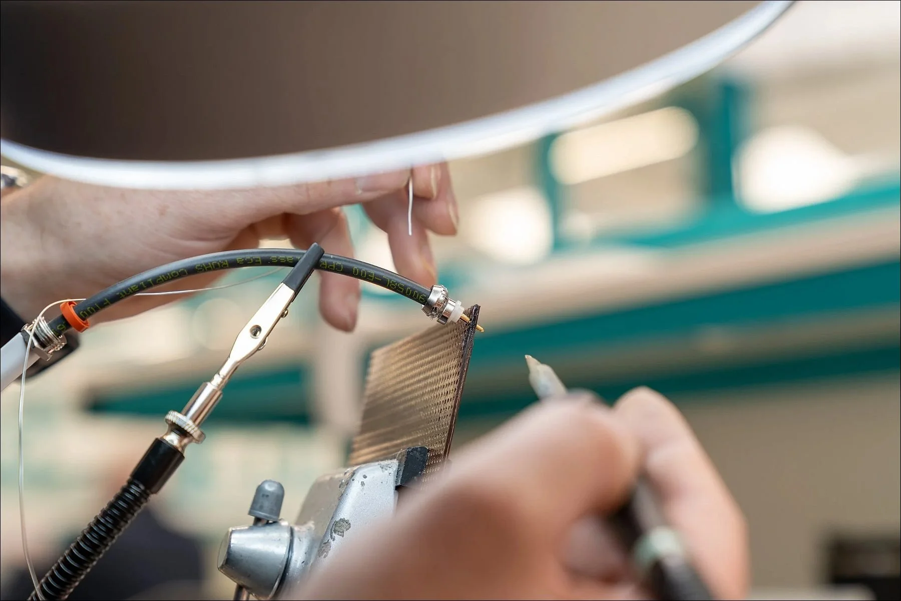 Close-up of hands soldering and connecting electrical wires using clamps under a magnifying glass at a workstation.