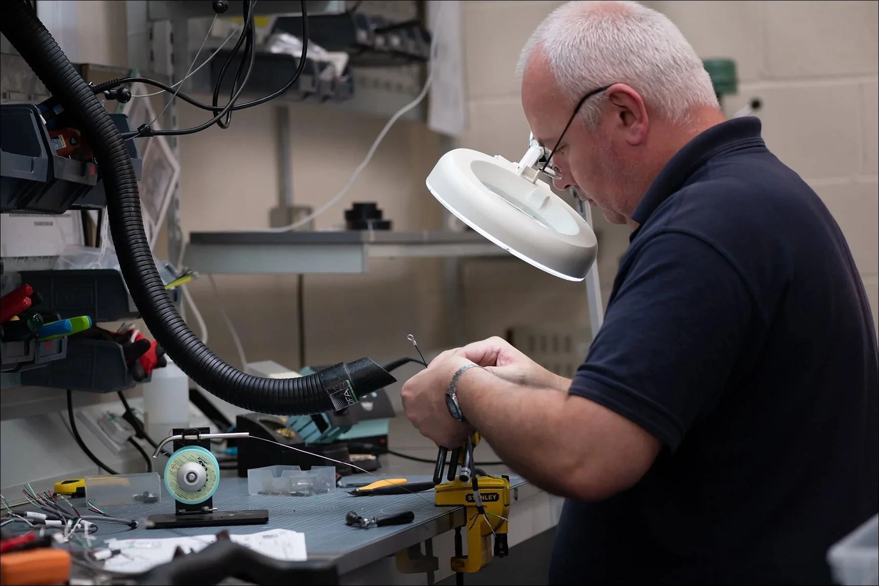 Technician inspecting and assembling large wiring harnesses under a magnifying lamp at a workstation.