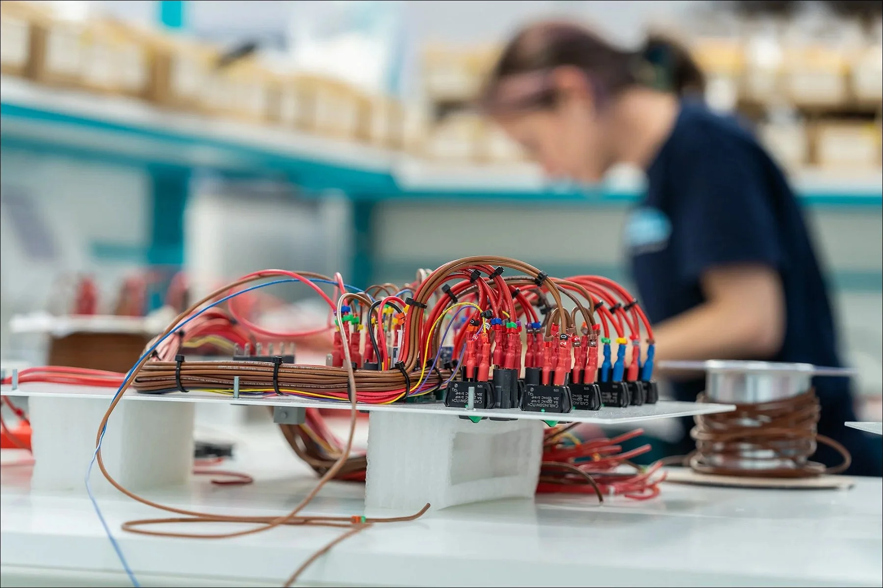 Close-up of red electrical cables wound around small spools with connectors, with workers blurred in the background.