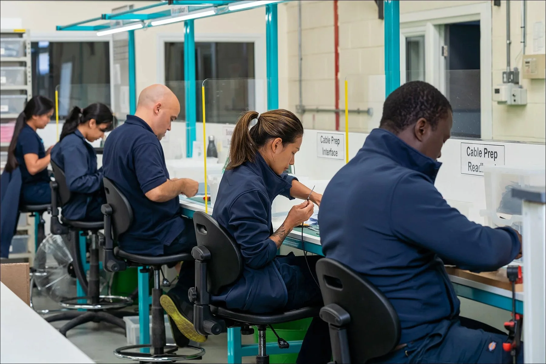 Multiple industrial workers seated at a production line assembling components on a factory floor.