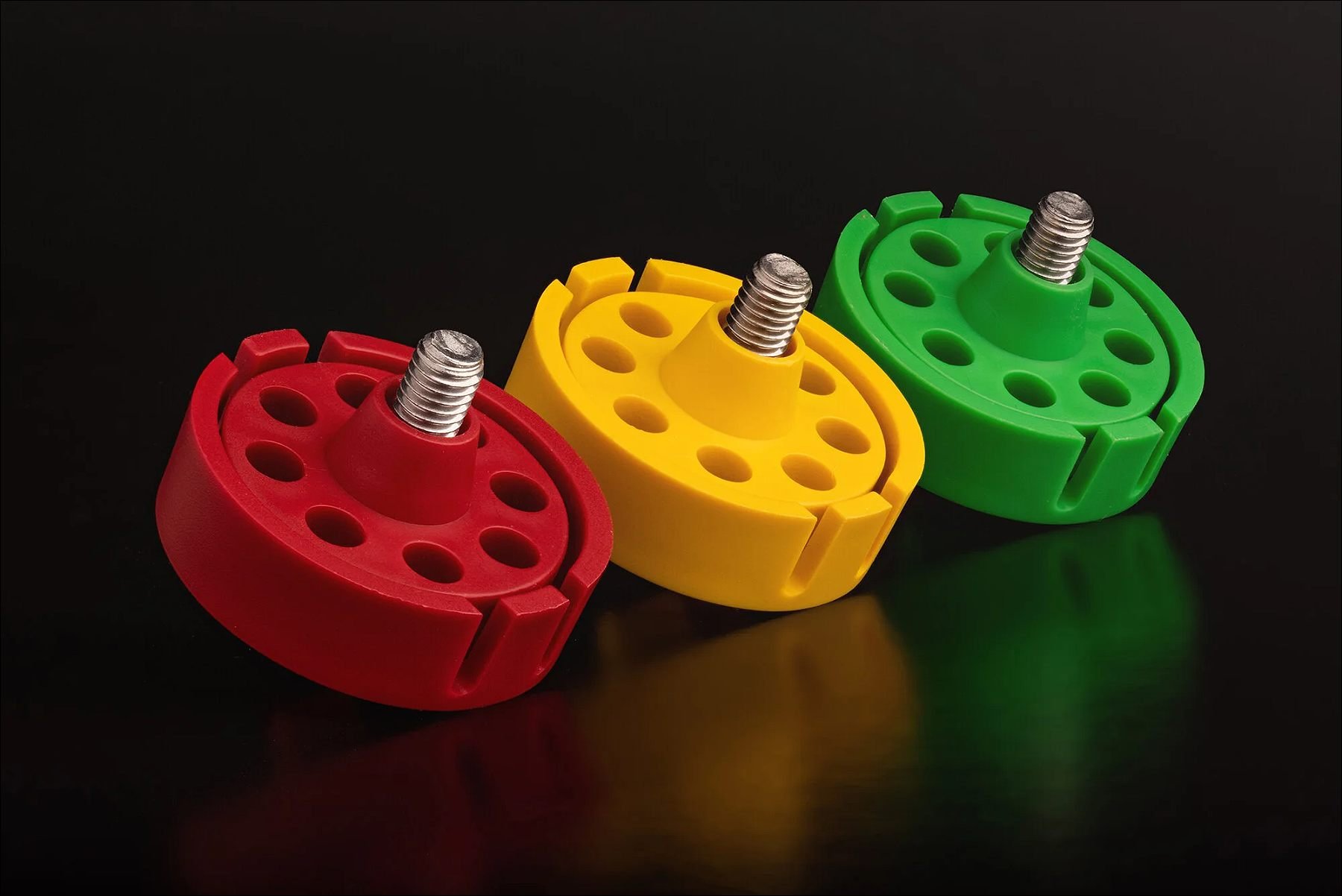 Three round mechanical components in red, yellow, and green with bolts inserted, arranged on a reflective surface against a dark background