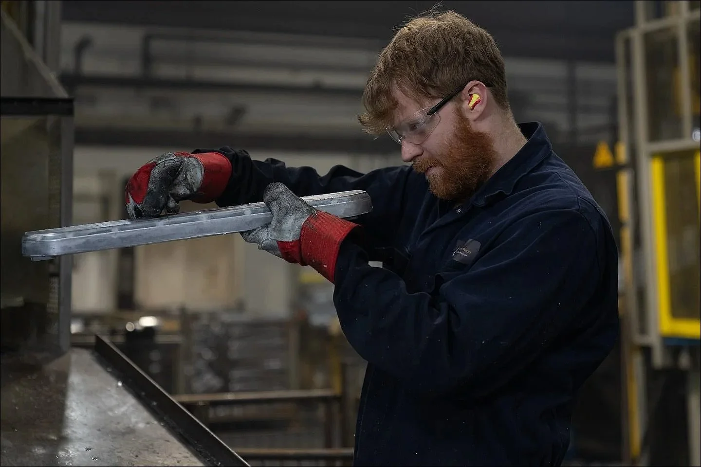 Worker wearing protective gloves and goggles using a power tool to cut or grind a metal pipe in a workshop.
