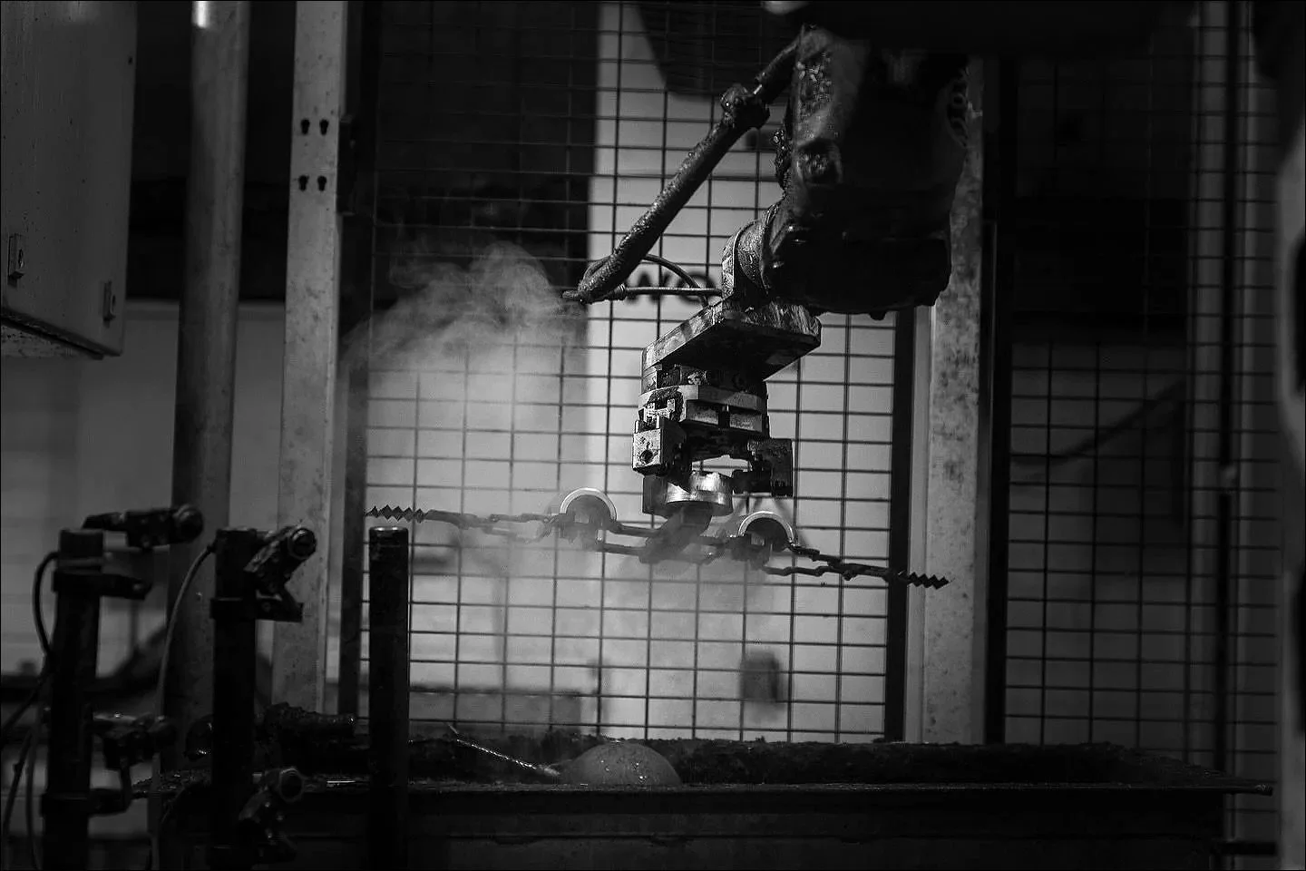 Black and white image of automated welding machinery operating behind a safety cage with smoke and sparks.