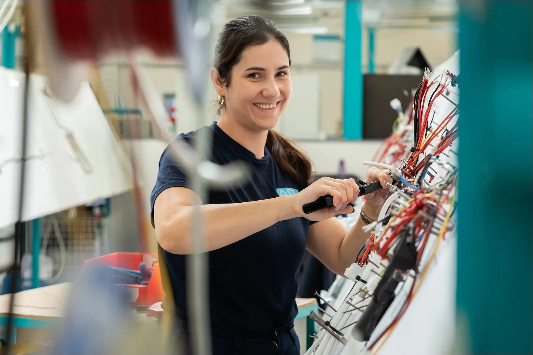Smiling female technician assembling wires on a harness board in a manufacturing facility.