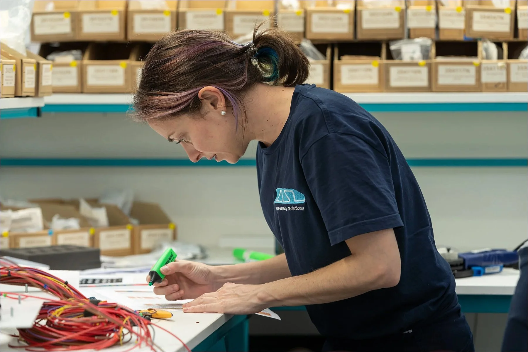 Worker concentrating on assembling red cables at a workstation with shelves of boxes in the background.