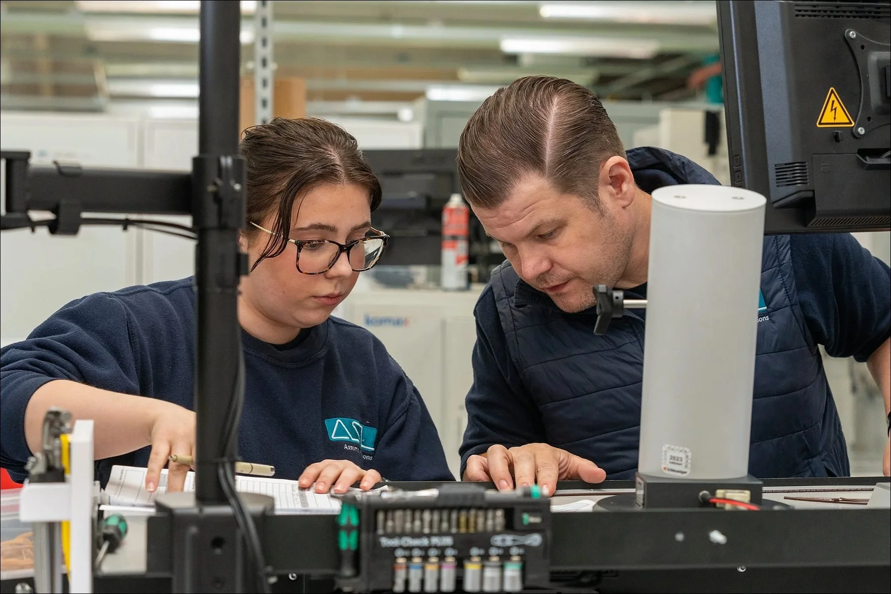 Two technicians collaborating at a workstation while examining and assembling electronic circuit components in a factory environment.