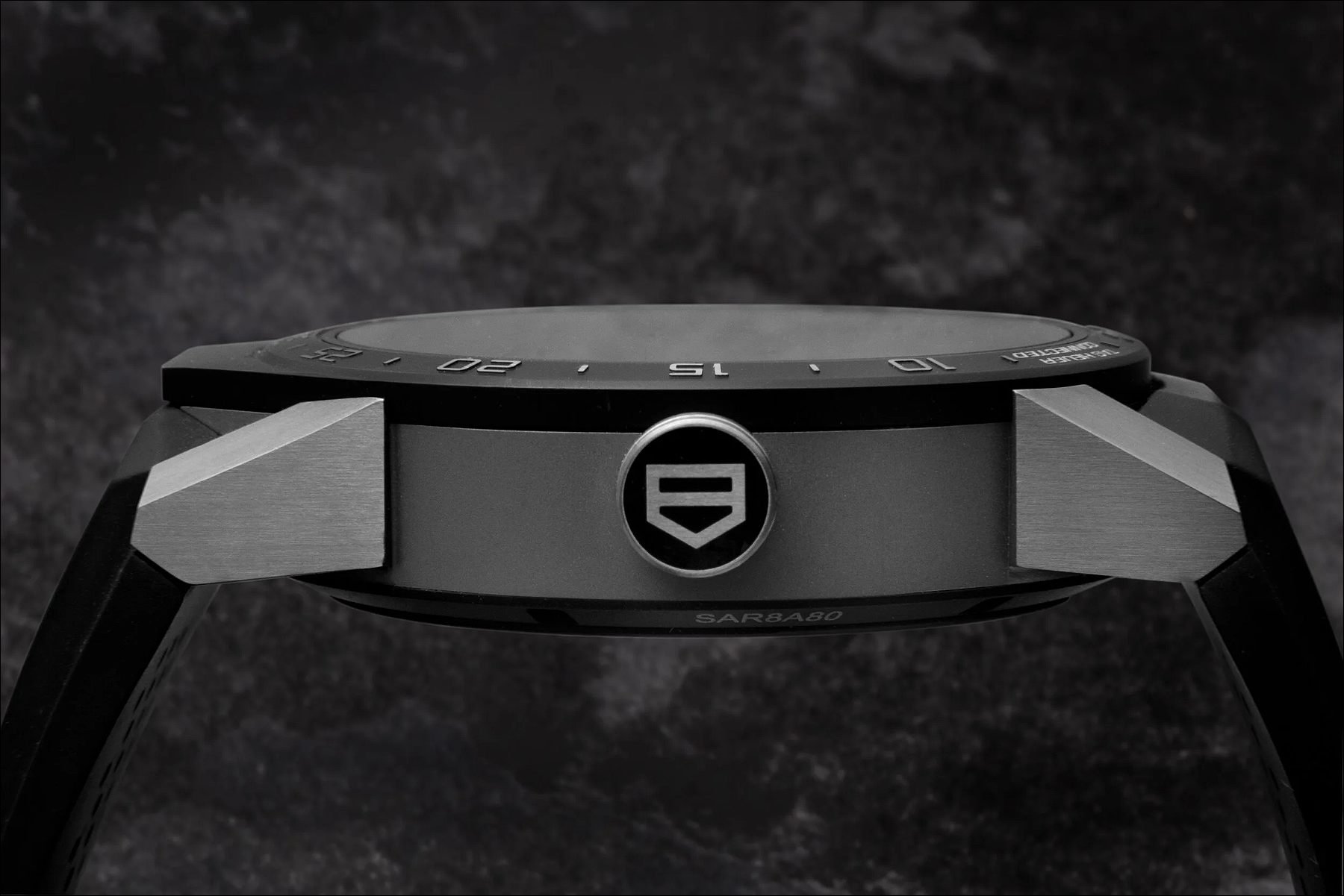 Side view of a black wristwatch with a logo on the case against a dark textured background