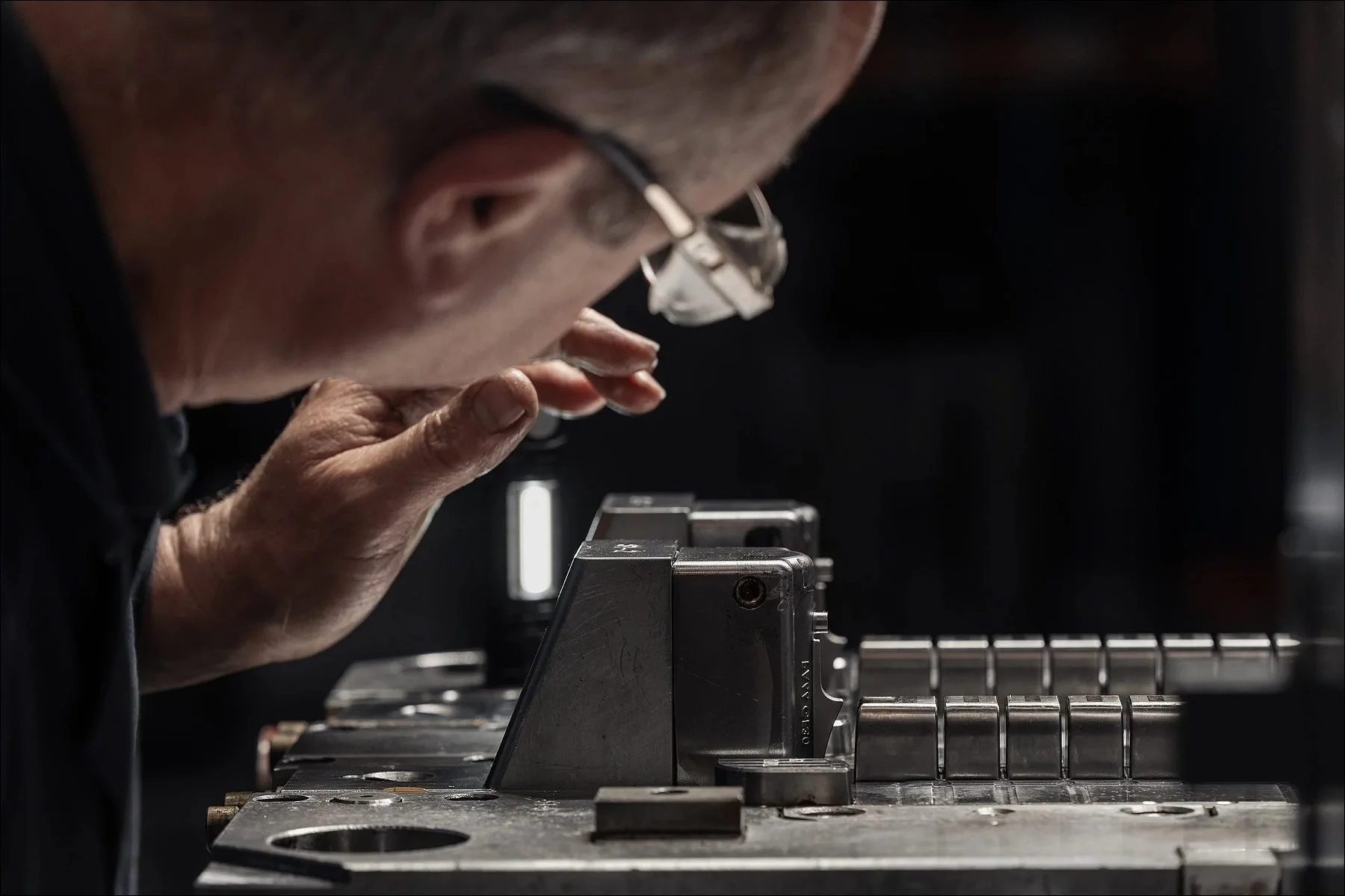 Worker wearing glasses focused on operating a bench vise to secure a component at a manufacturing workstation.