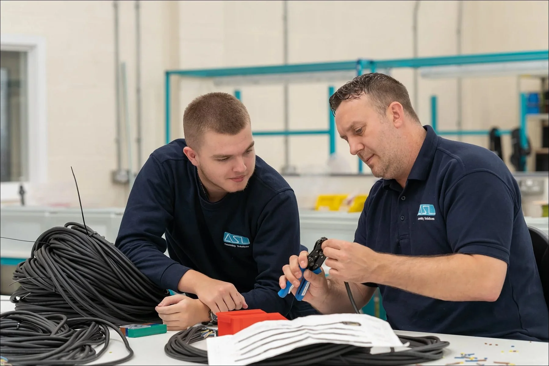 Two factory workers collaborating at a workstation with coils of cables and documents.