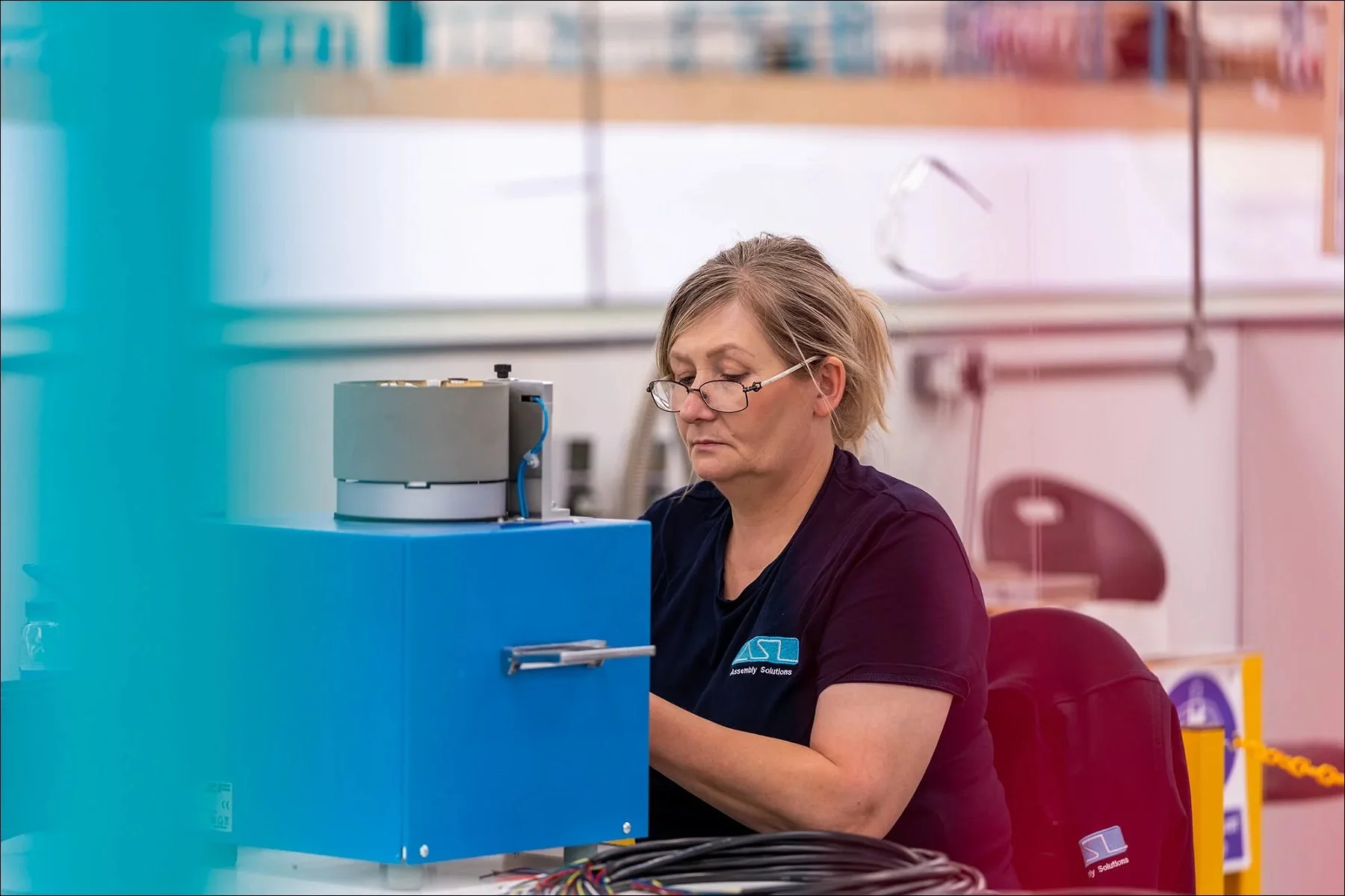 Factory worker operating equipment behind a transparent blue safety shield at a workstation.