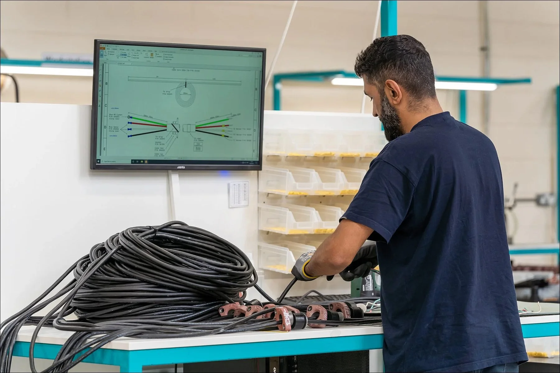 Worker standing at a workstation assembling or organizing black cable harnesses while referencing instructions on a monitor.
