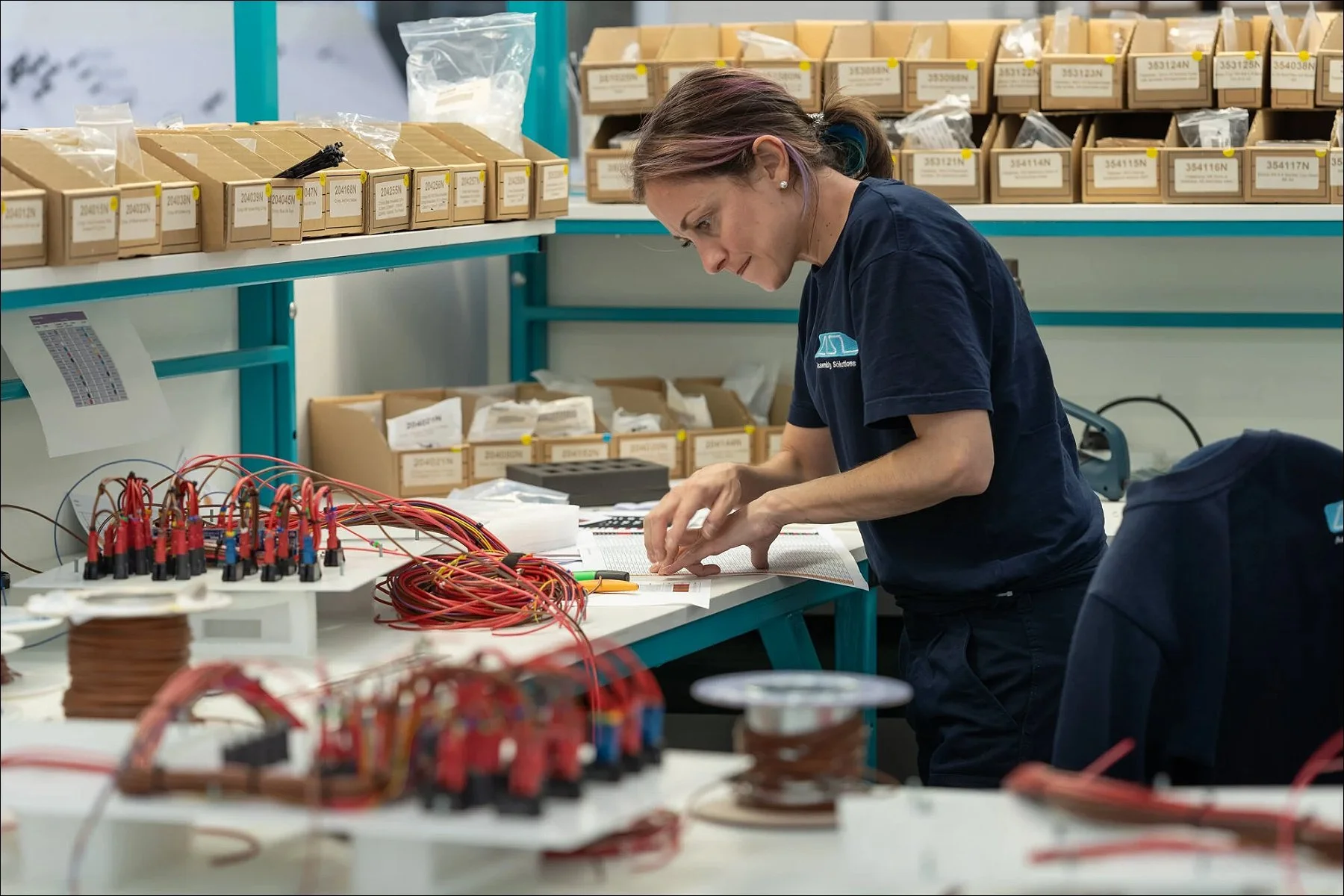 Factory worker managing spools of red cable at a workstation surrounded by storage boxes.