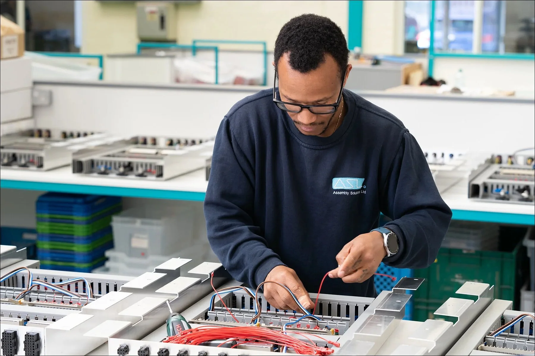 Technician assembling electronic components on a long workstation in a factory environment.