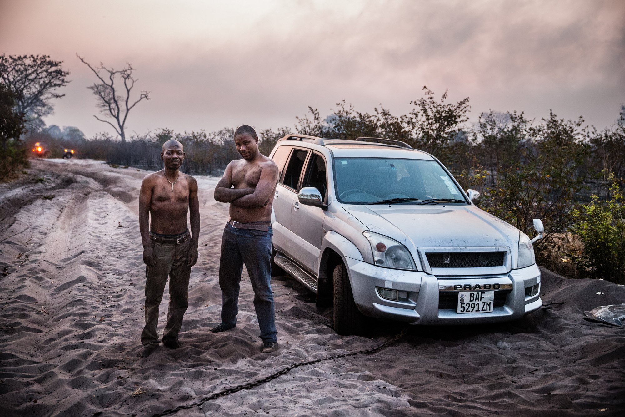 Two shirtless men standing next to a white Toyota Prado SUV on a sandy dirt road. The background features sparse trees and a smoky sky.
