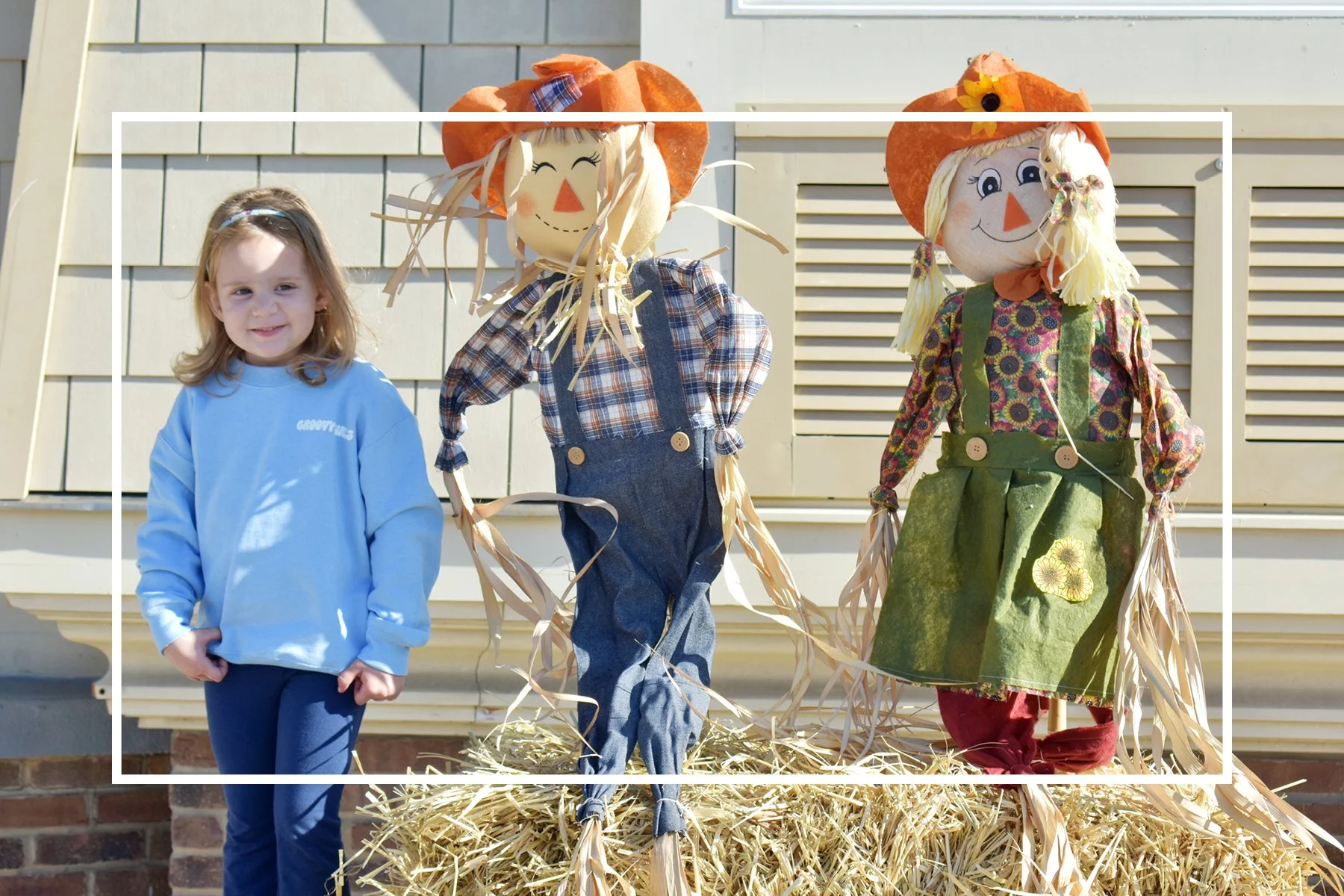 young girl in blue sweatshirt standing next to two scarecrows